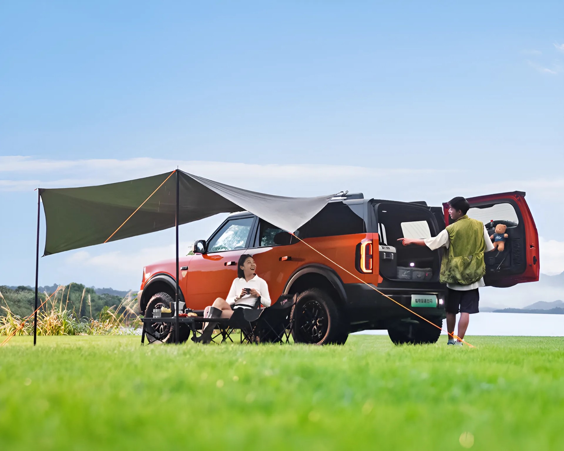 Orange SUV with black roof and tires parked on grass with a gray canopy, camping chairs, and two people.