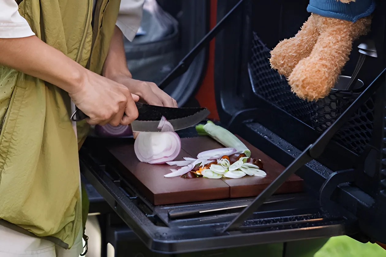 Person slicing a red onion on a wooden cutting board placed on a vehicle's tailgate with a stuffed animal hanging nearby.