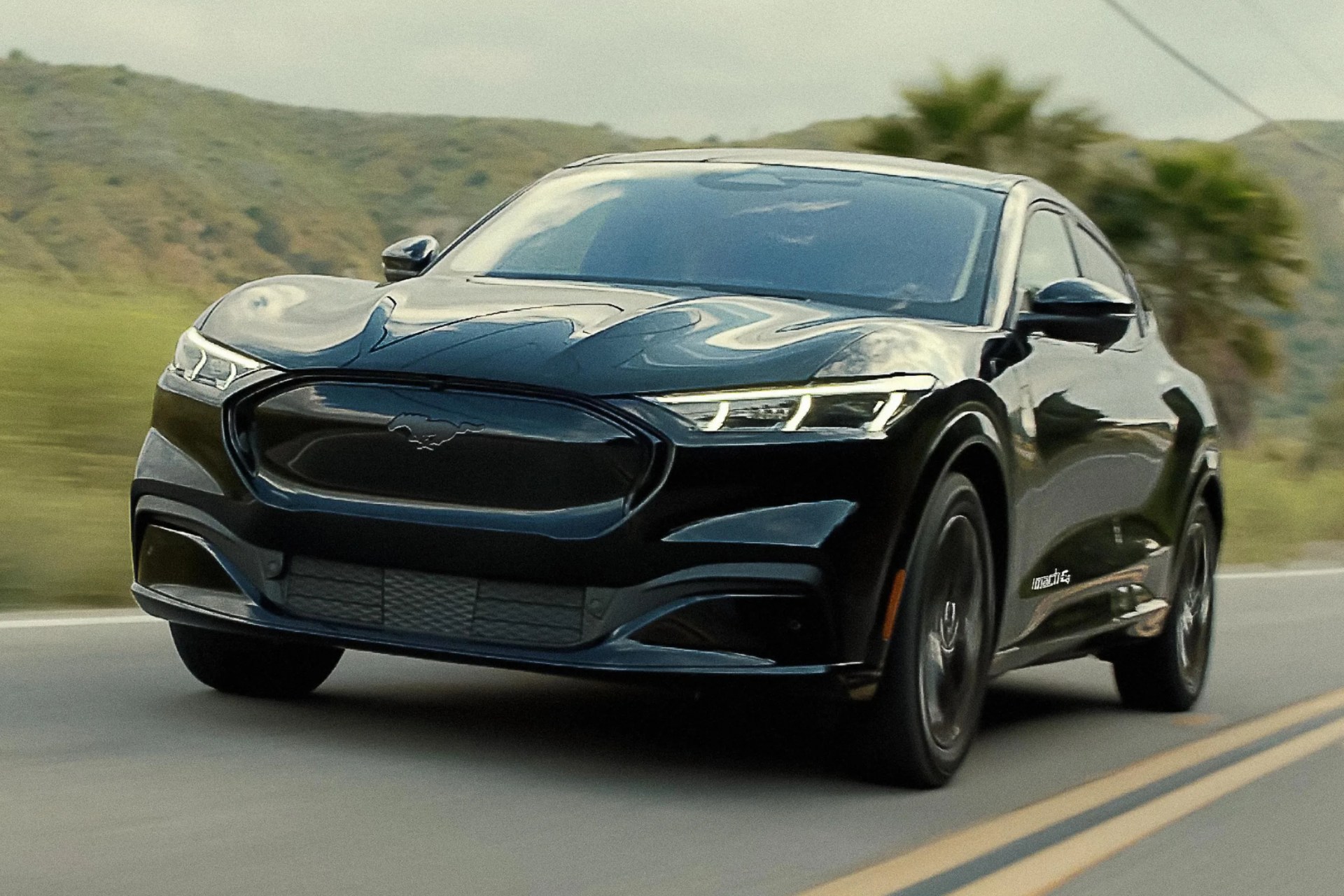 Black Ford Mustang Mach-E electric SUV driving on a road with hills and palm trees in the background.