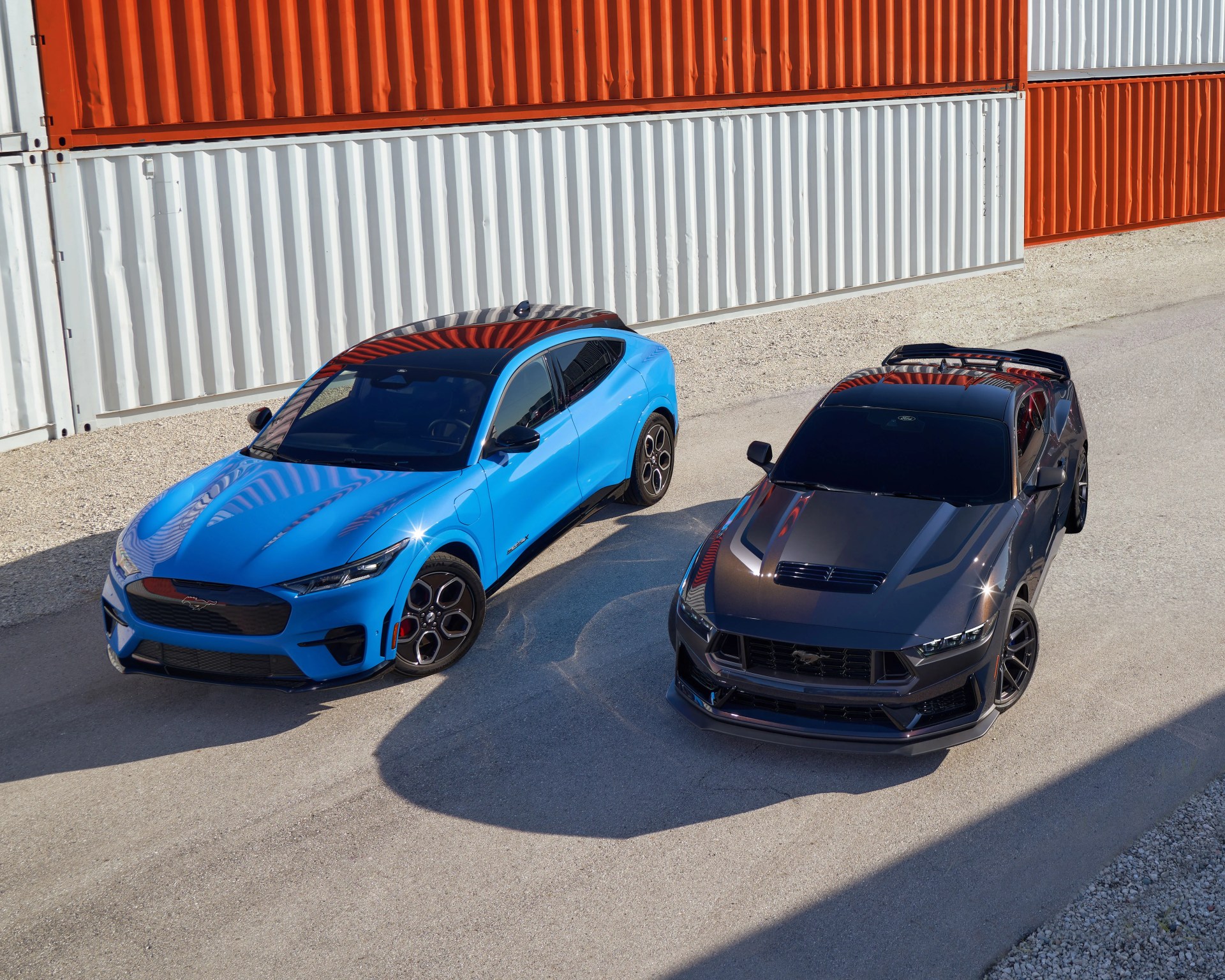 Blue Ford Mustang Mach-E electric SUV and dark gray Ford Mustang sports car parked side by side on a paved surface with orange and white shipping containers in the background.