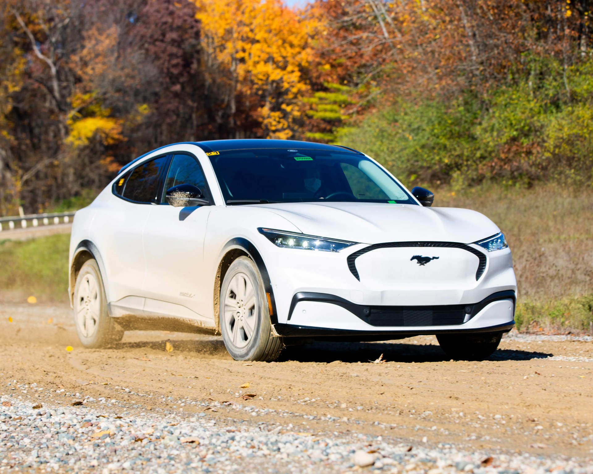 White Ford Mustang Mach-E electric SUV driving on a dirt road with autumn trees in the background.
