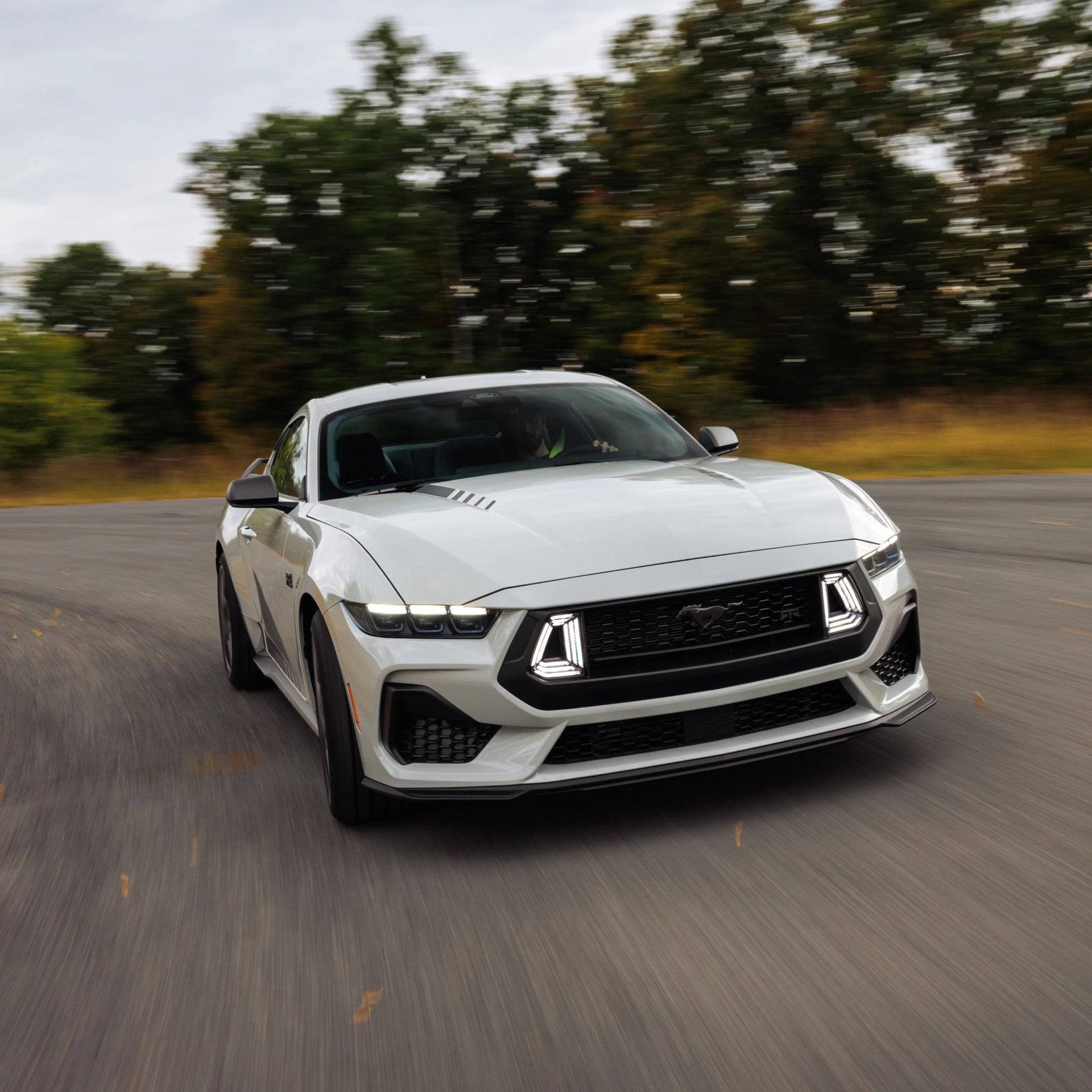 White Ford Mustang GT with LED headlights driving on a curved road with blurred trees in the background.