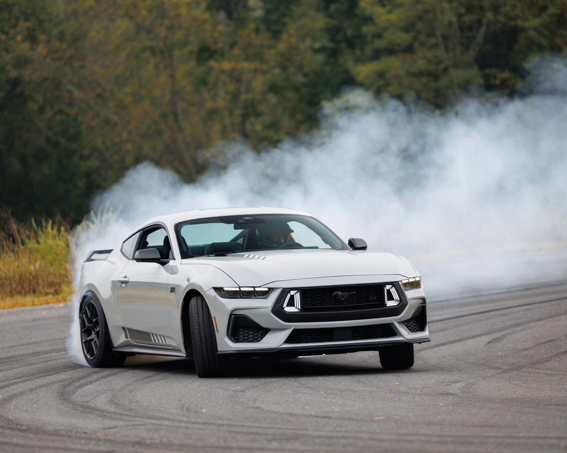 White Ford Mustang RTR drifting on a paved road with smoke trailing from the rear tires.