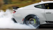 White sports car performing a burnout with smoke around the rear tire and visible yellow Brembo brake caliper.