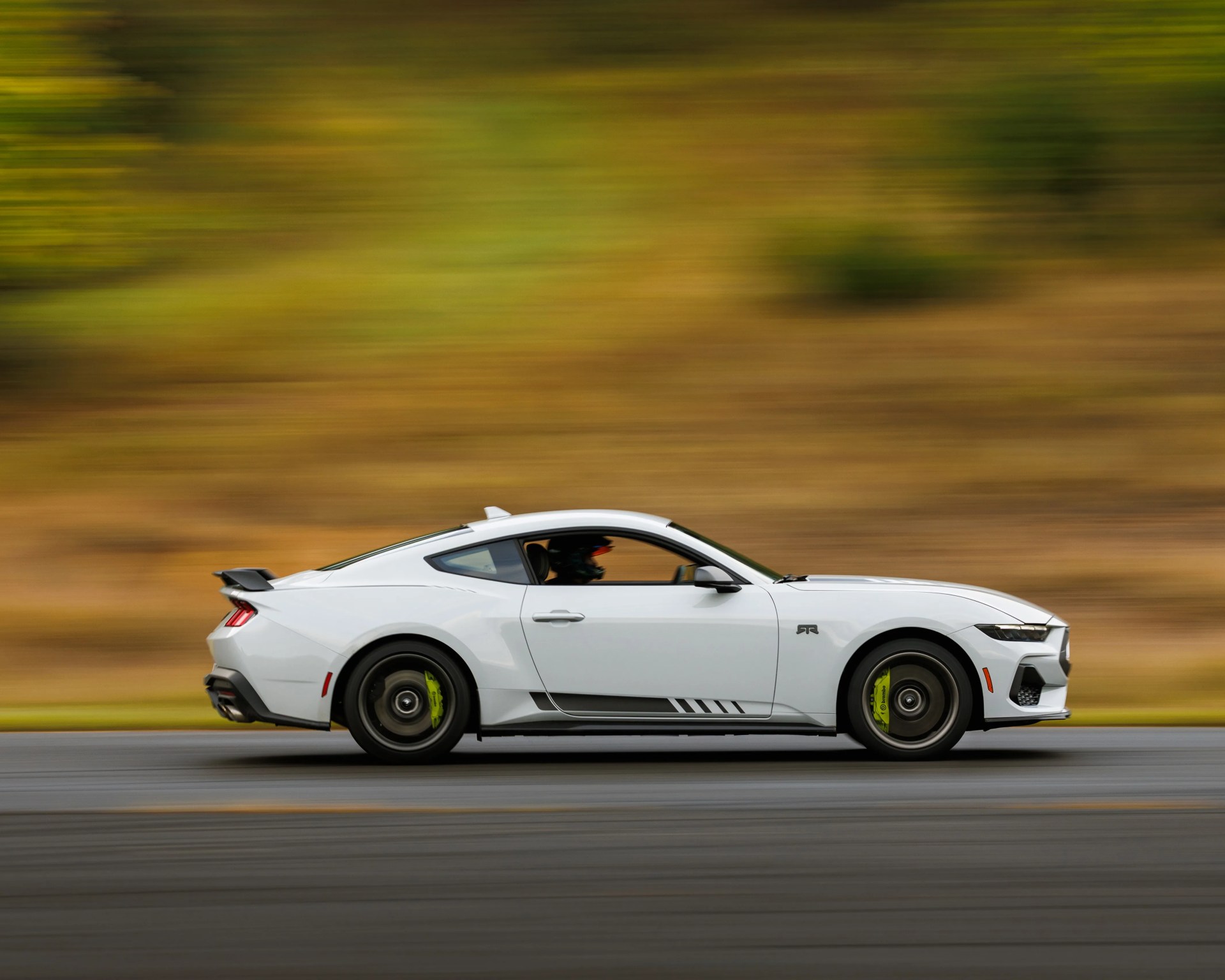 White sports car with black wheels and green brake calipers driving on a road with blurred autumn foliage background.