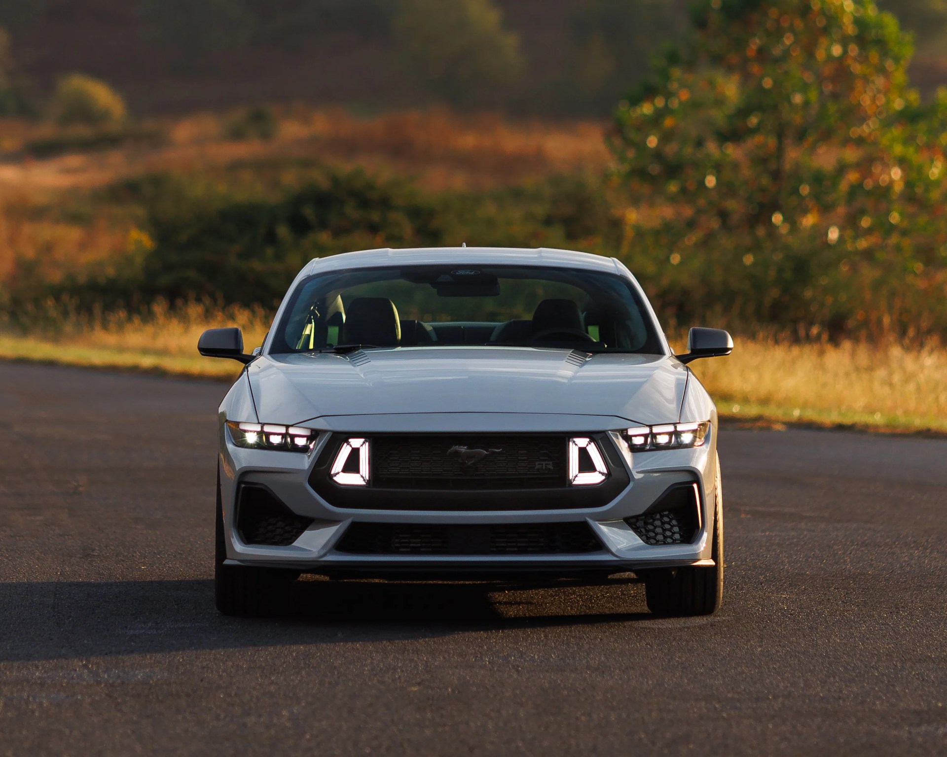 White Ford Mustang RTR with illuminated headlights and grille lights on a paved road with blurred greenery in the background.