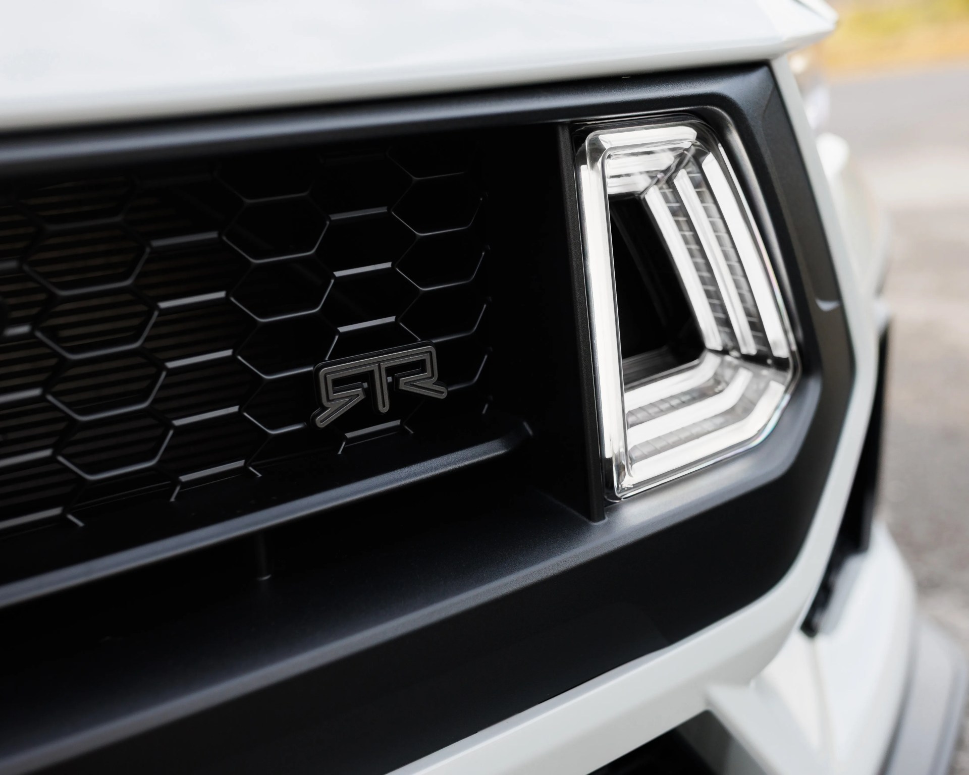 Close-up of a white vehicle's front grille with a black honeycomb pattern and an "RTR" emblem next to a uniquely shaped LED headlight.