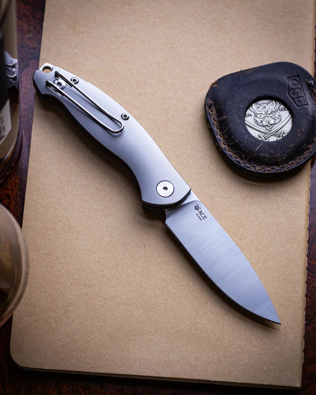 Silver folding knife with clip and "ACE" logo on blade, resting on a tan notebook next to a black leather coin pouch.