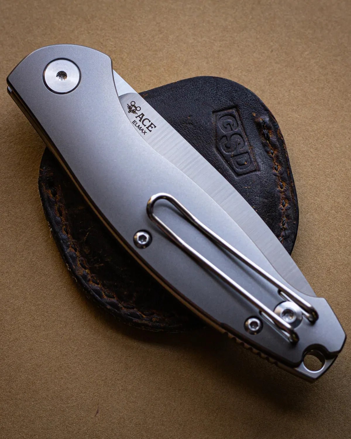 Closed silver folding knife with a metal clip resting on a black leather GSD sheath on a brown surface.