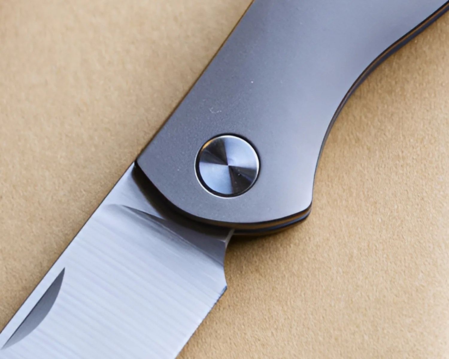 Close-up of a folding knife with a matte gray handle and a polished stainless steel blade on a beige surface.