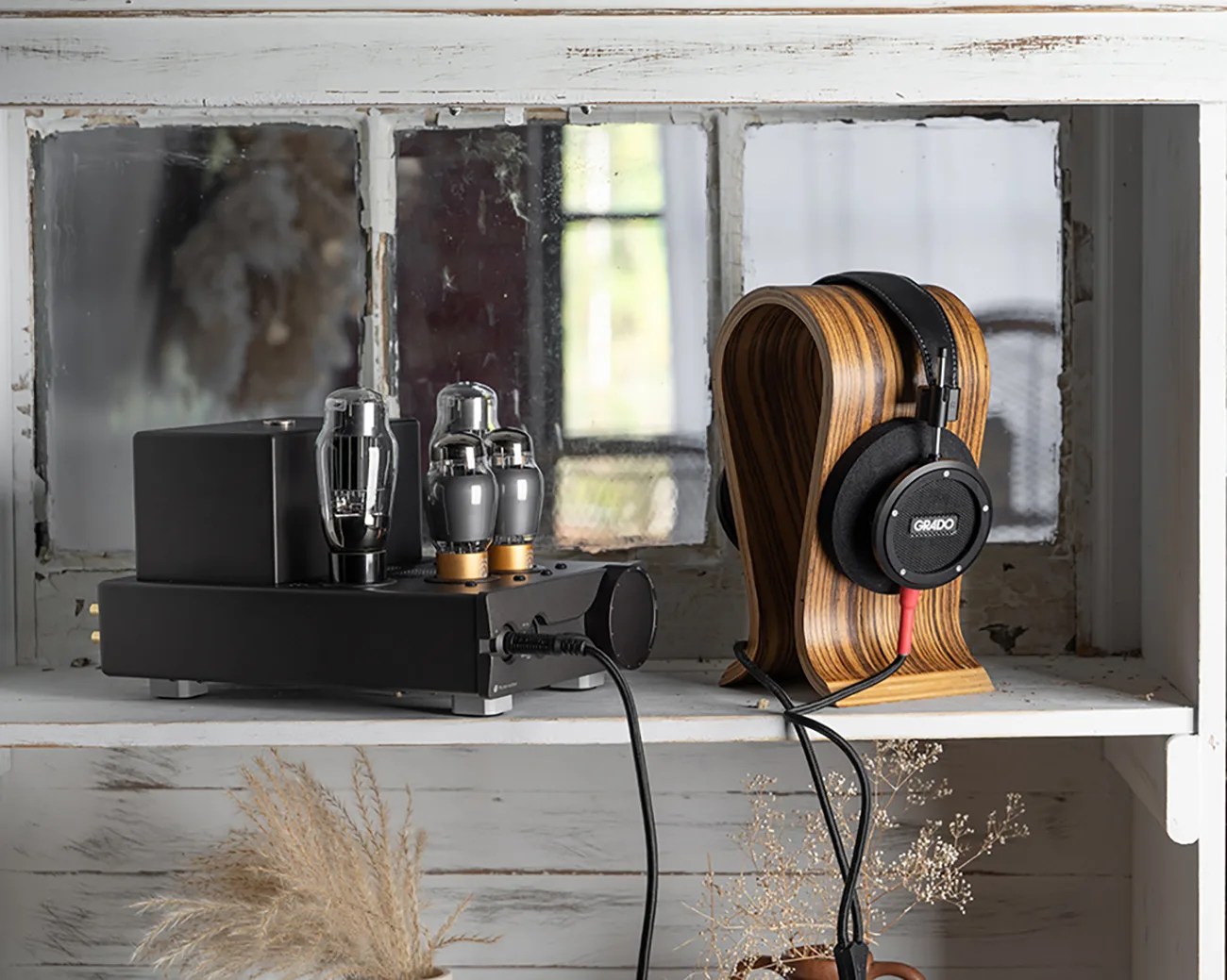 Black tube headphone amplifier with three glass vacuum tubes next to black Grado headphones on a curved wooden stand on a white shelf.