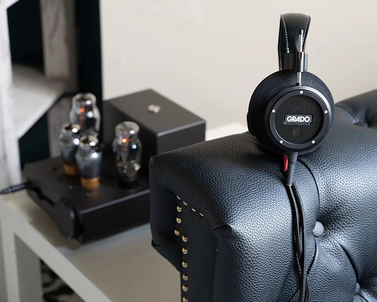 Black Grado headphones resting on the arm of a black leather tufted sofa with a tube amplifier in the background.