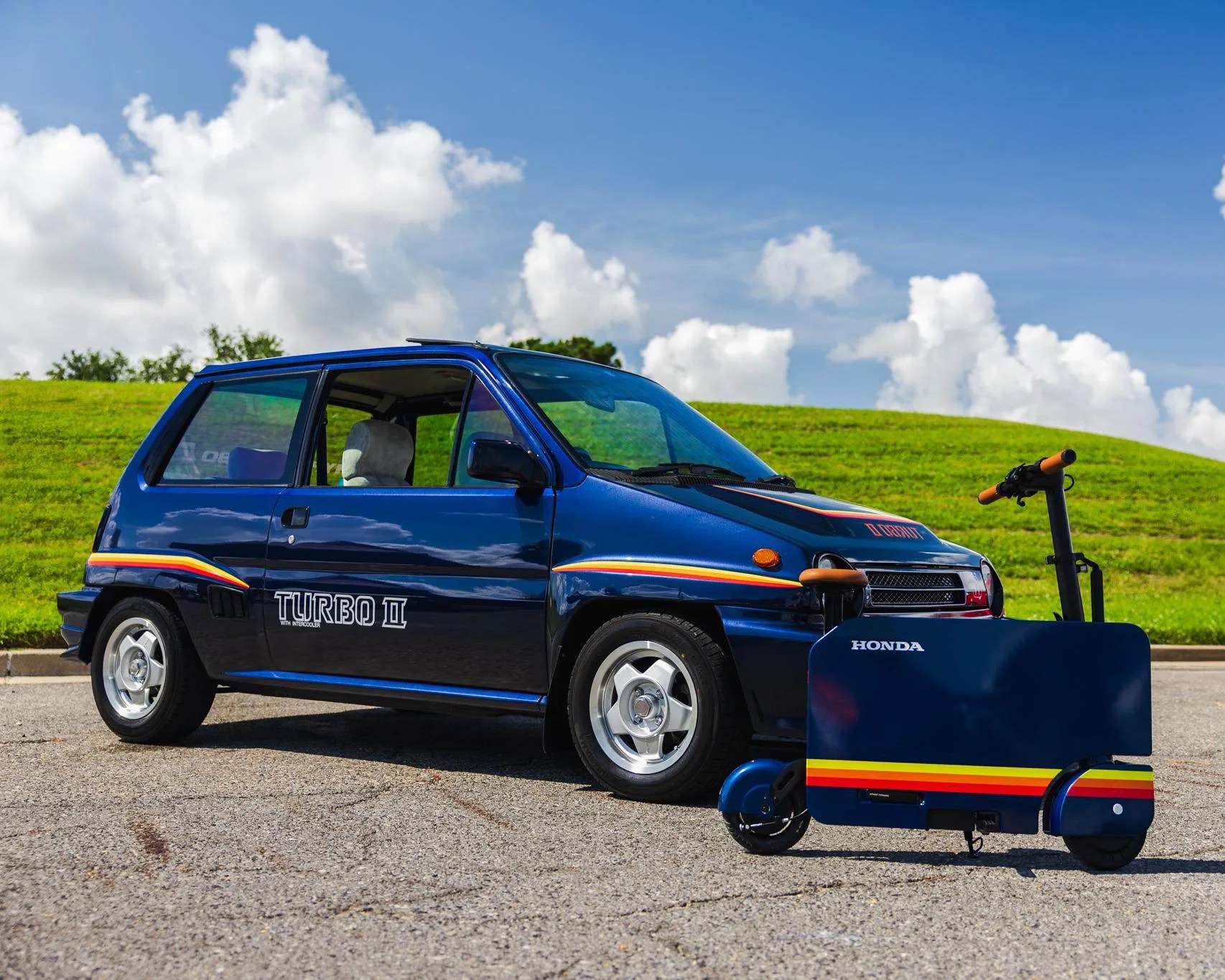 Blue Honda Turbo II hatchback car with orange and yellow stripes parked next to a matching blue Honda electric scooter with orange handlebars.