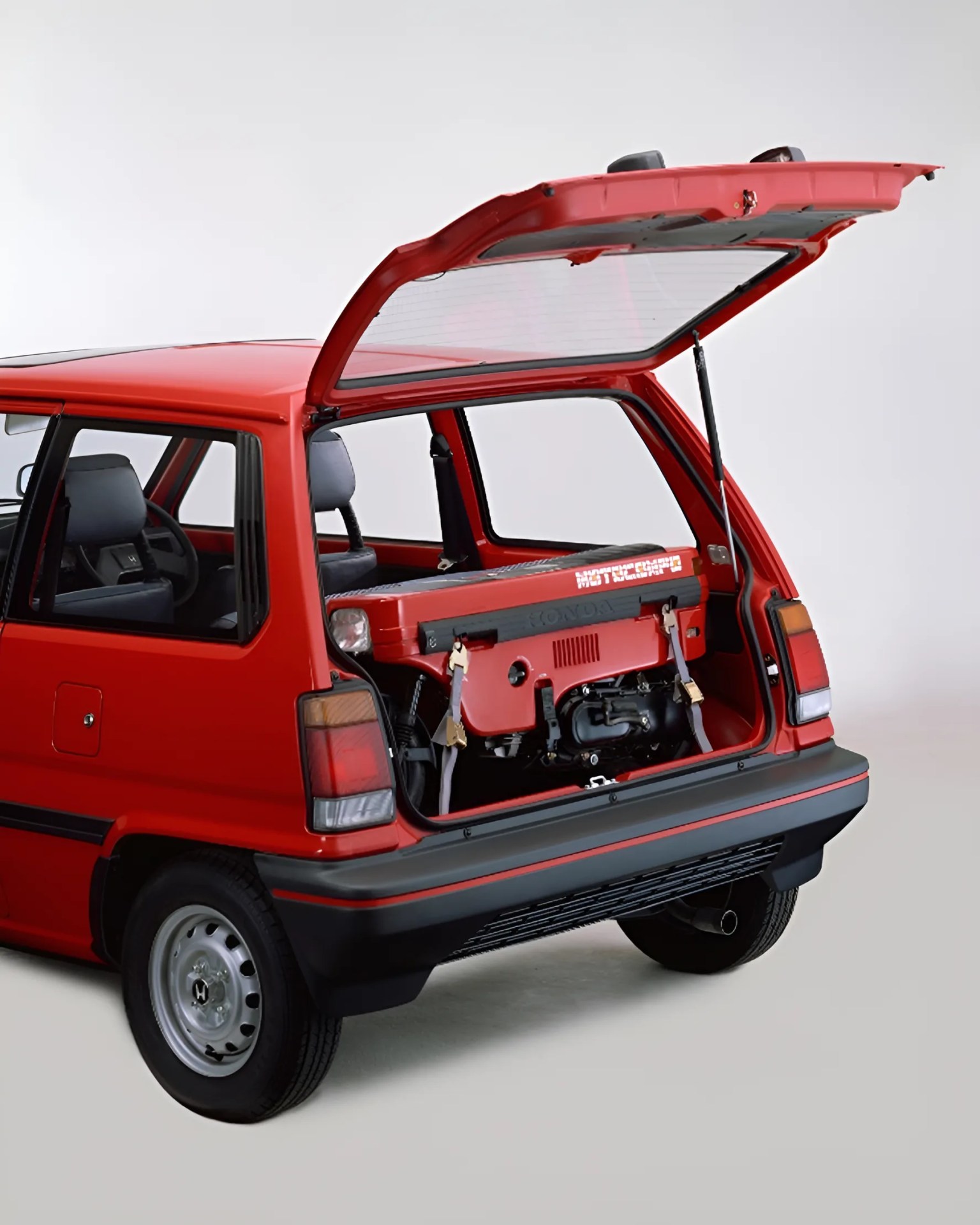 Red compact hatchback car with rear hatch open, showing a red Honda Motocompo foldable scooter inside.