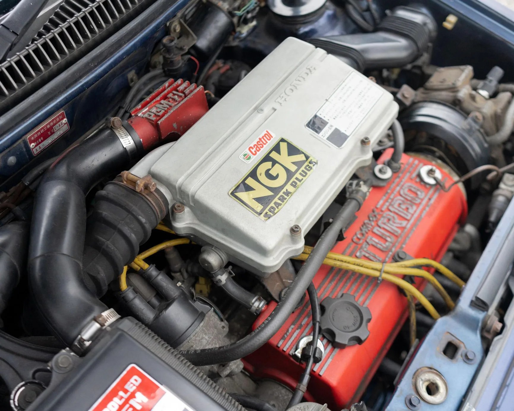Close-up of a Honda engine with a gray cover labeled NGK spark plugs and a red valve cover marked "turbo" with yellow ignition wires.