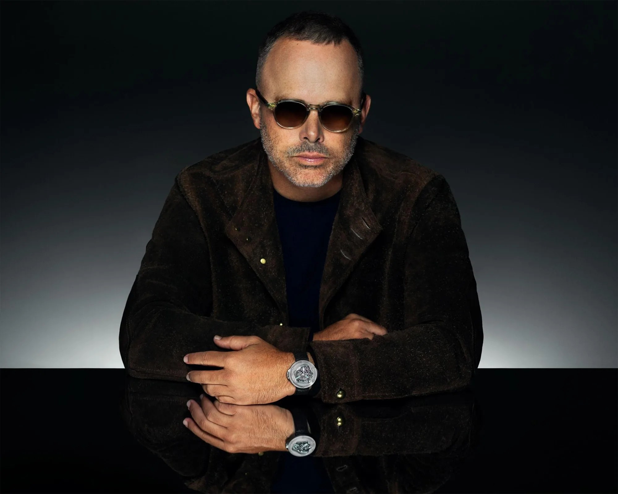 Man wearing round sunglasses and a dark brown suede jacket, resting arms on a reflective black surface showing a silver watch.