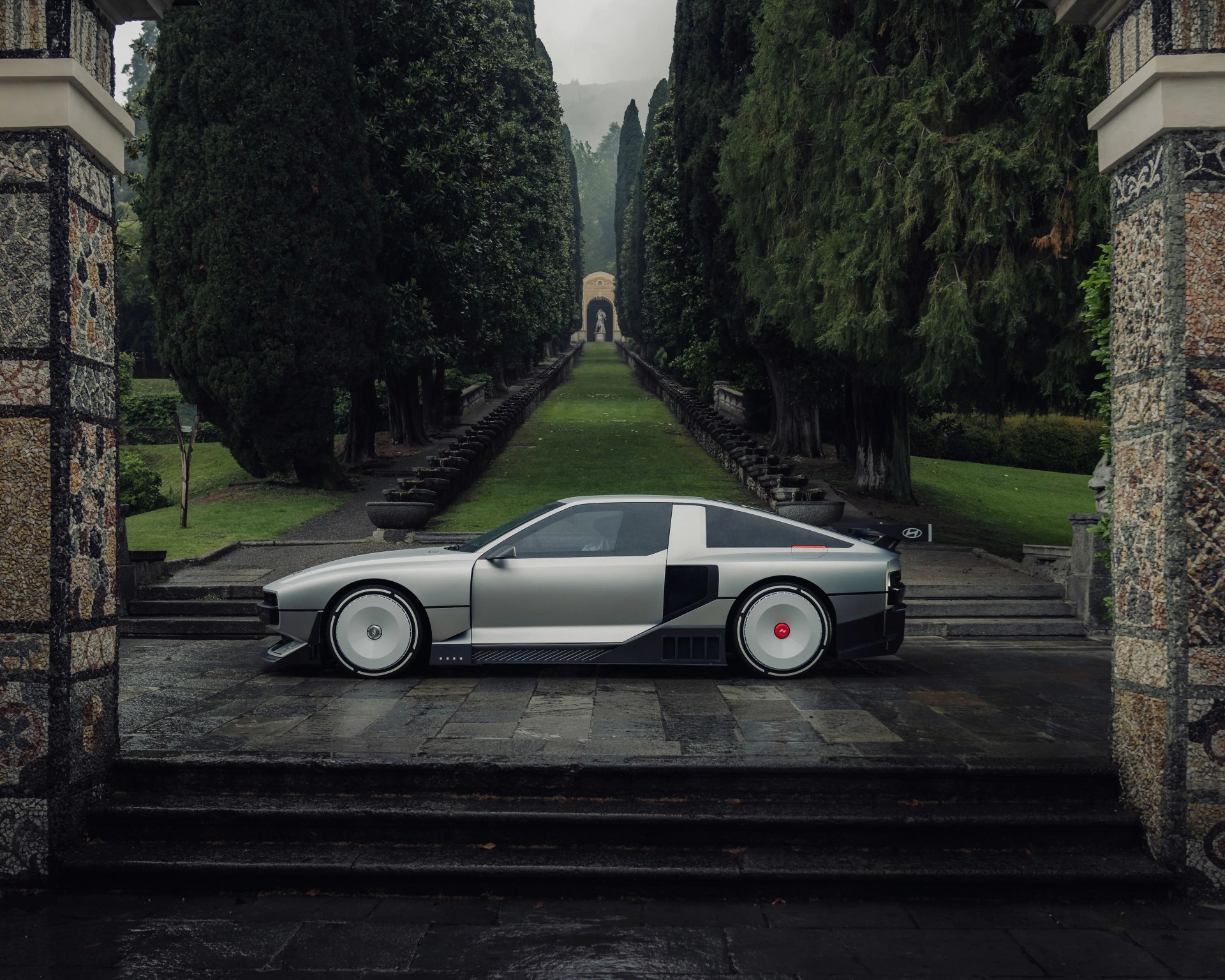Silver futuristic sports car with white disc wheels parked on stone pavement between tall trees.