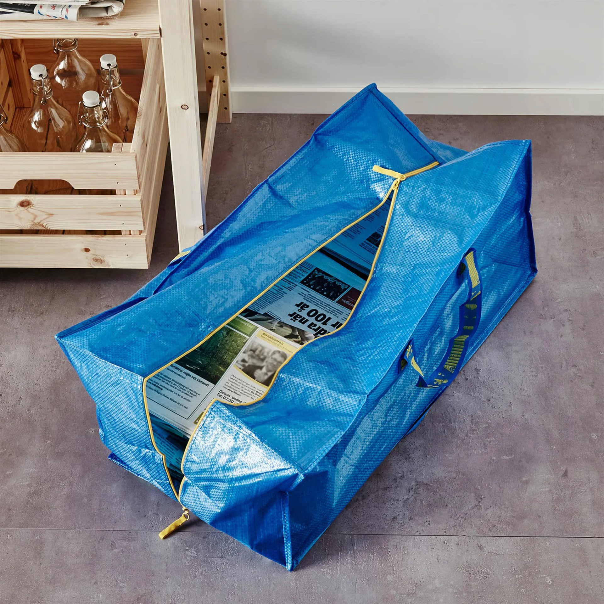 Large blue IKEA storage bag with yellow zipper partially open, filled with newspapers, on a gray floor next to wooden crates with glass bottles.