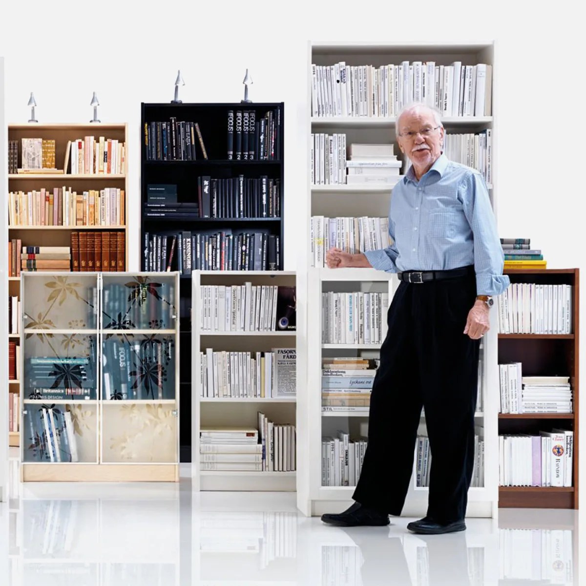 Older man in a blue shirt and black pants standing next to white bookshelves filled with white books.