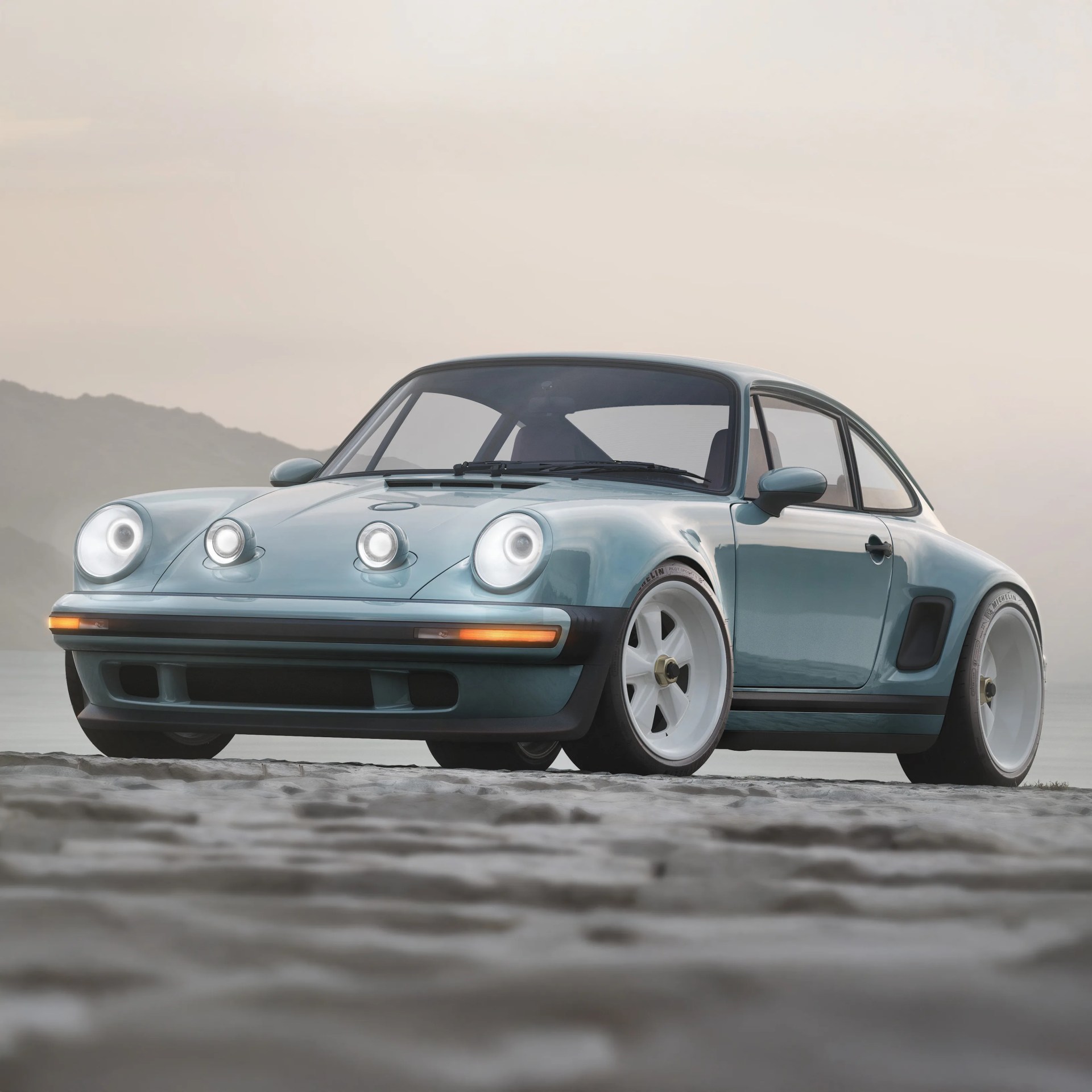 Light blue vintage sports car with wide white wheels and additional front hood lights on a sandy surface.