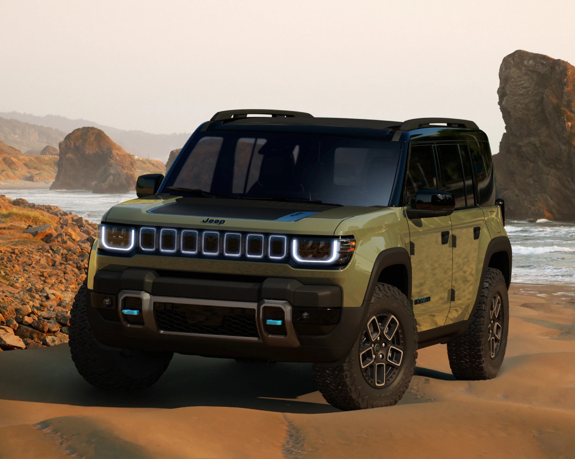 Olive green Jeep SUV with black roof and illuminated grille parked on a sandy beach near rocky cliffs.