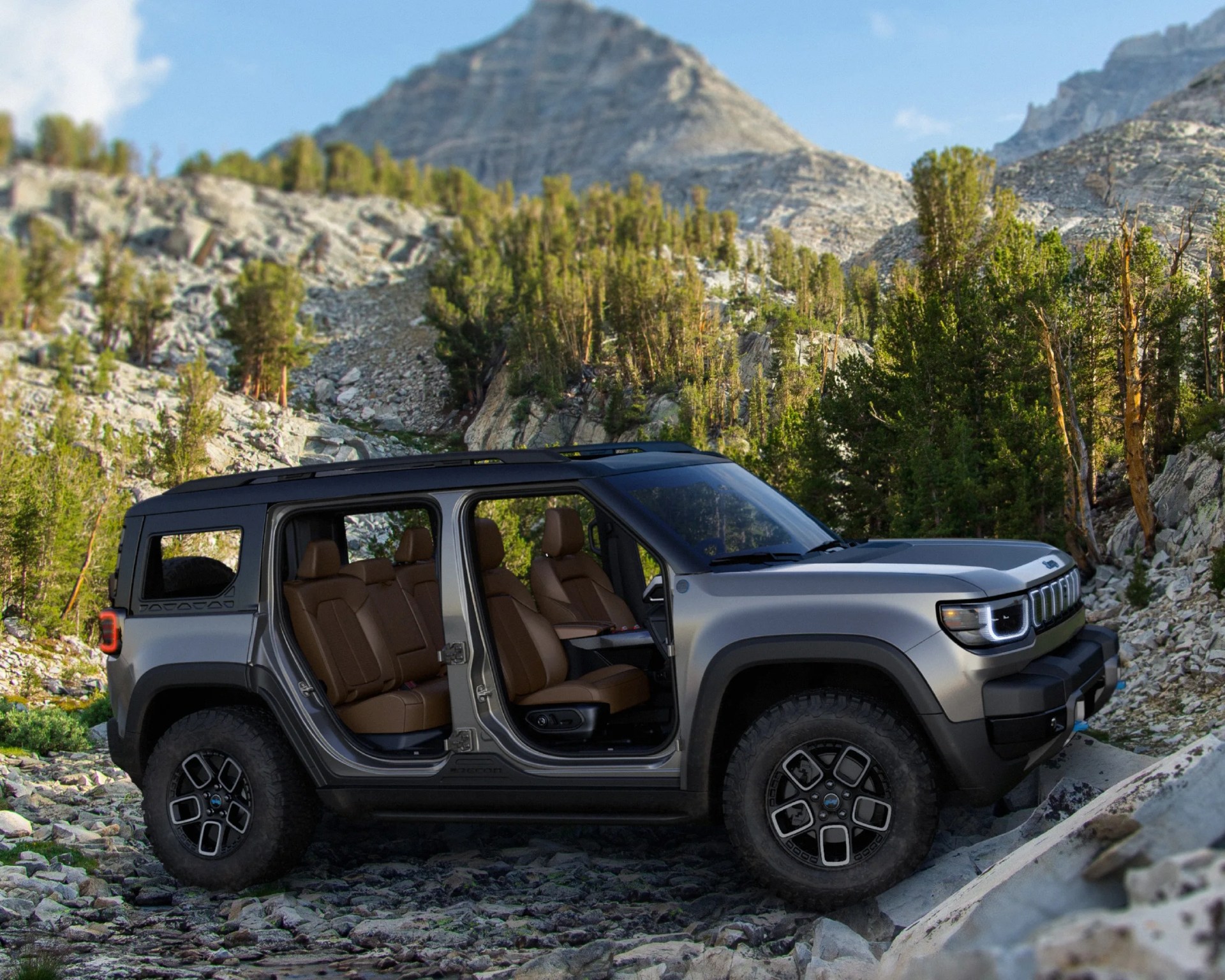 Silver off-road SUV with open doors showing brown leather seats parked on rocky terrain with trees and mountains in the background.