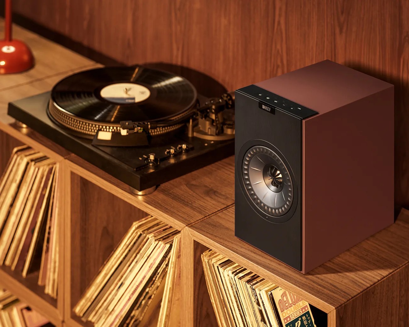 Brown KEF speaker with black front panel and silver driver on a wooden shelf next to a turntable and vinyl records.