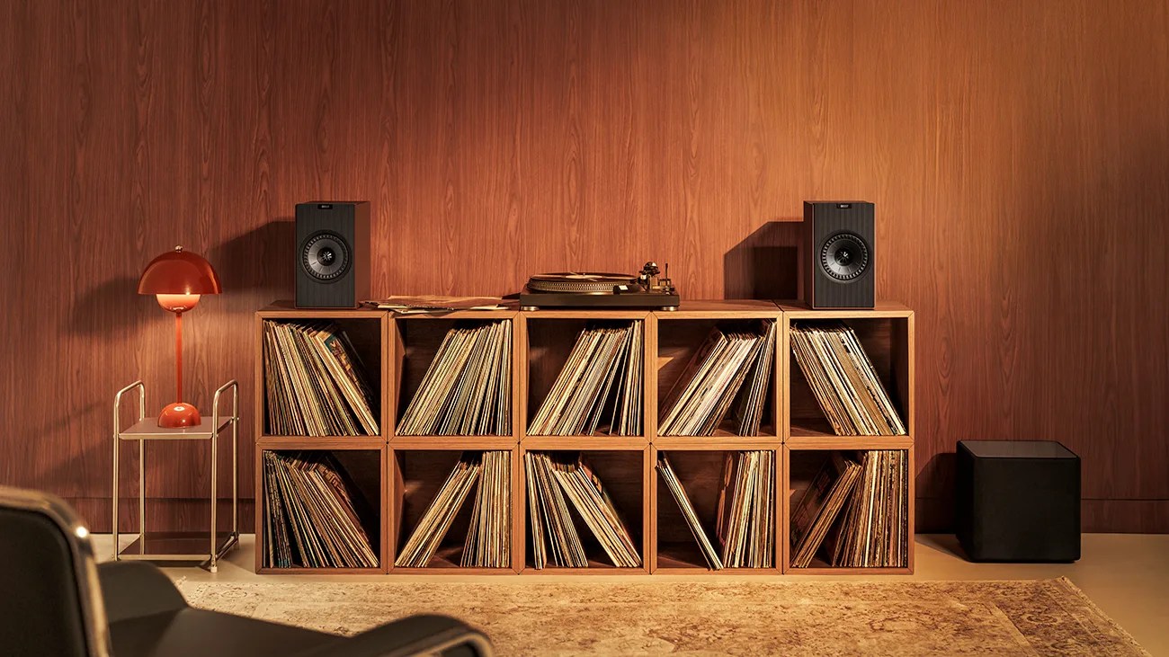 Wooden shelving unit filled with vinyl records, a turntable, two black speakers, and a red lamp on a side table against a wood-paneled wall.