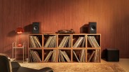 Wooden shelving unit filled with vinyl records, a turntable, two black speakers, and a red lamp on a side table against a wood-paneled wall.