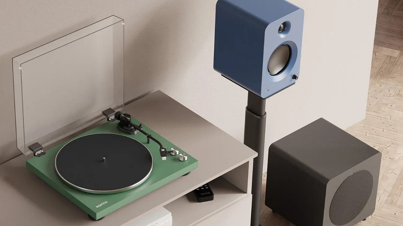 Green Kanto turntable with clear dust cover on a beige shelf next to a blue speaker on a stand and a black subwoofer on a wooden floor.