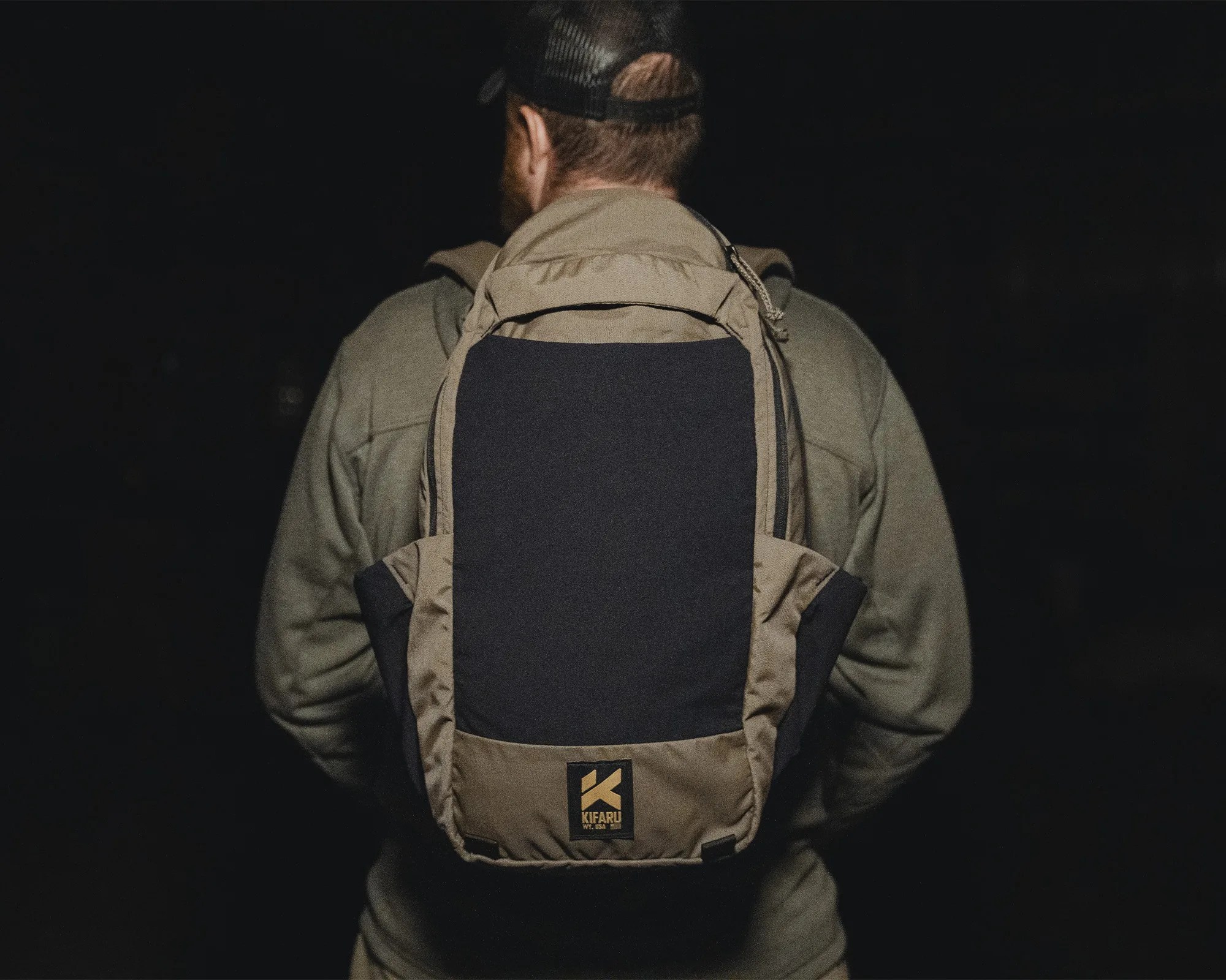 Tan and black Kifaru backpack worn by a person facing away against a dark background.