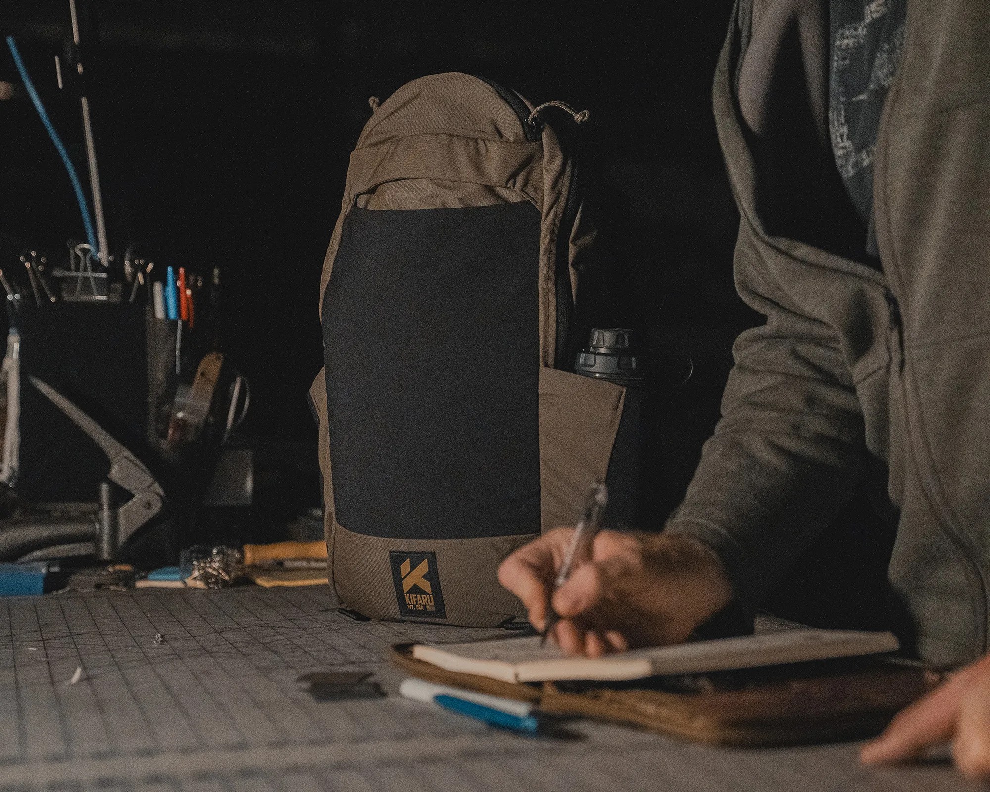 Brown and black Kifaru backpack with water bottle in side pocket on a workbench next to a person writing in a notebook.