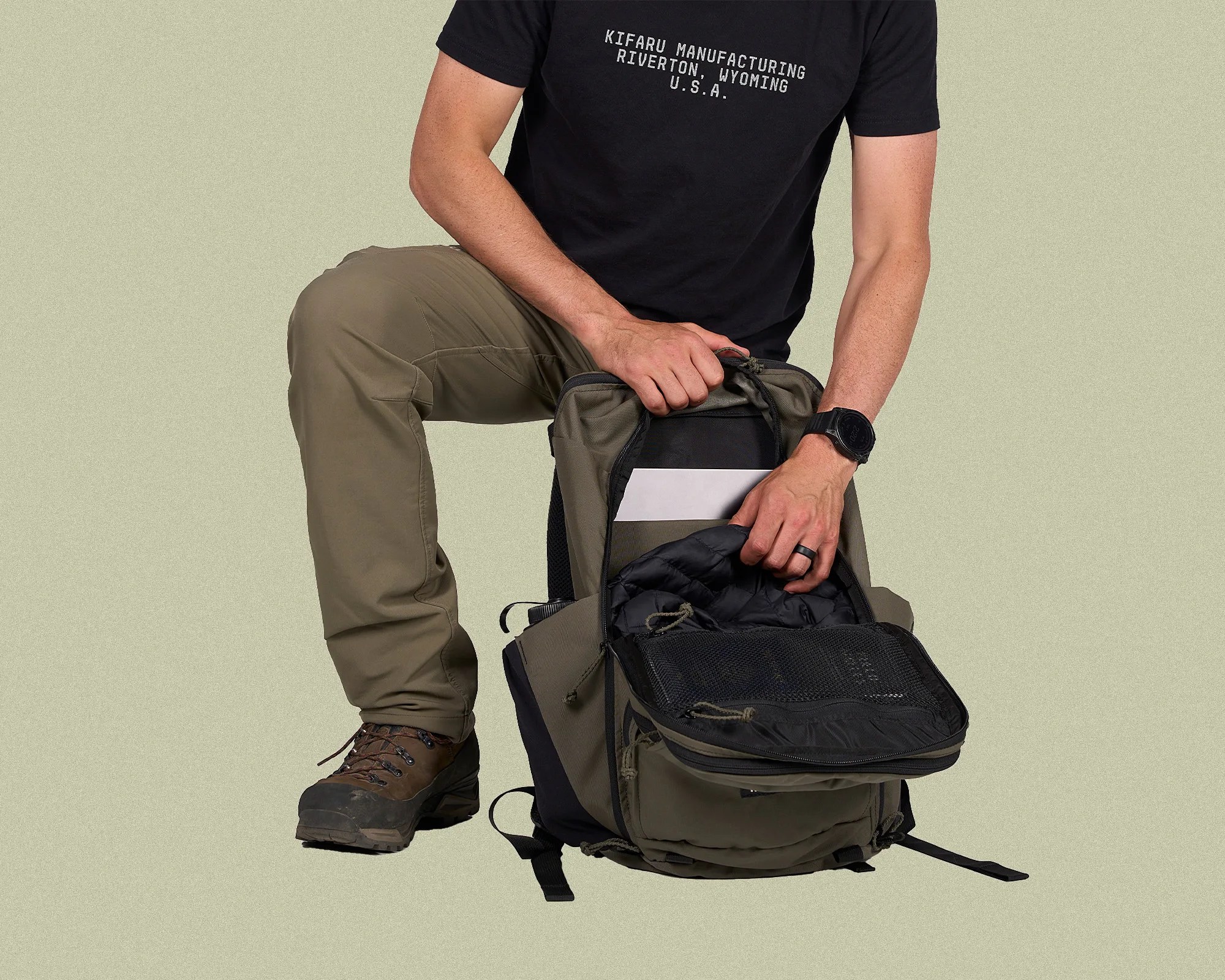Person wearing brown pants and hiking boots holding an open olive green and black backpack with mesh pocket.