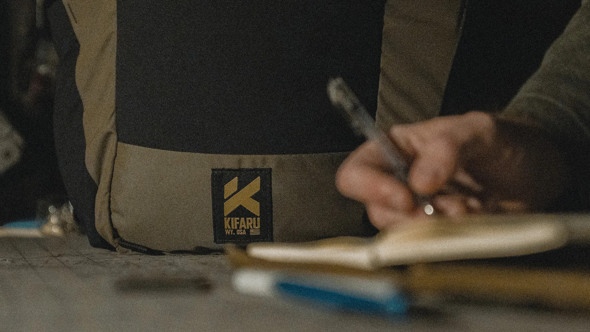 Close-up of a black and olive green Kifaru backpack with a person writing on a notepad in the foreground.