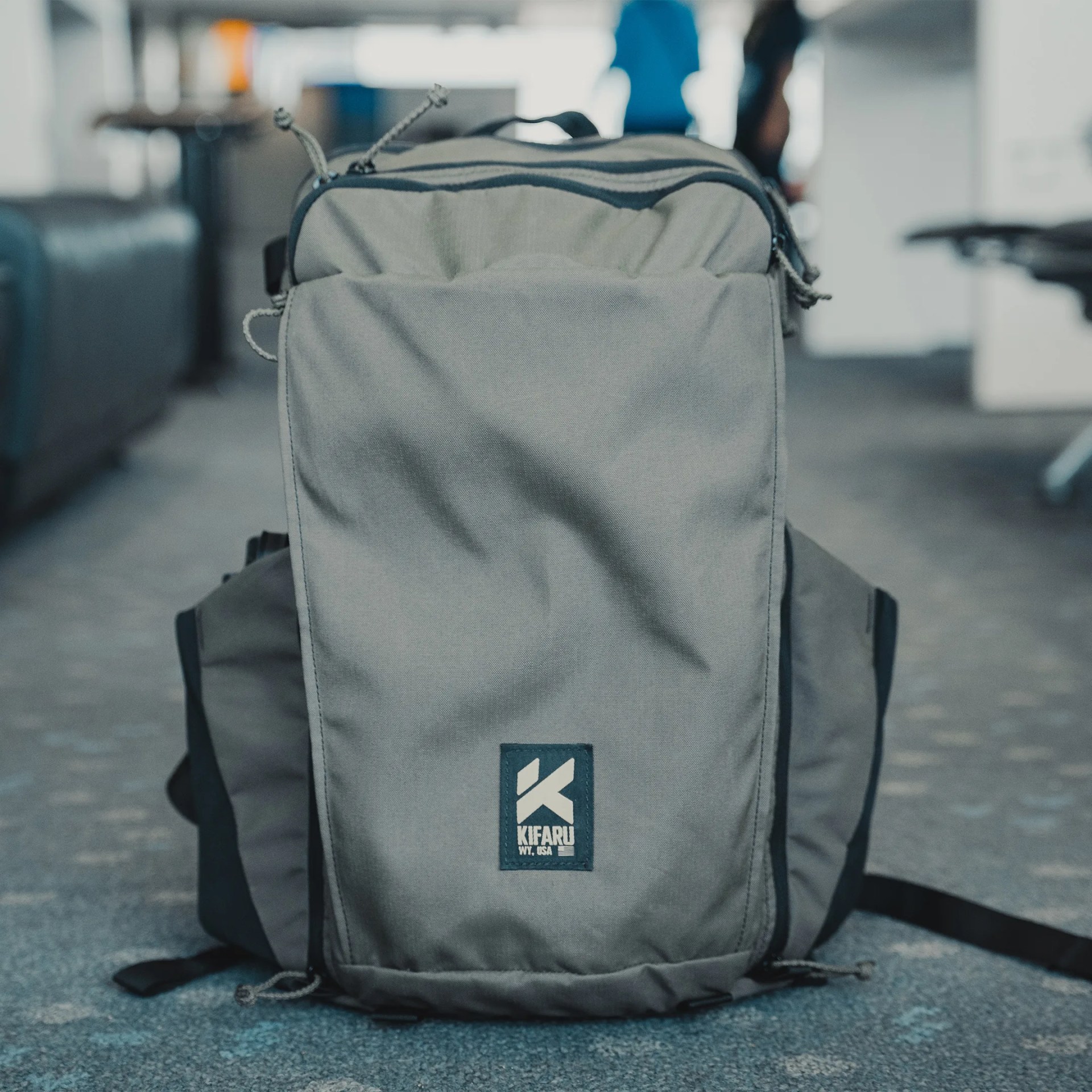 Gray Kifaru backpack with front logo patch on a carpeted floor in an indoor setting.