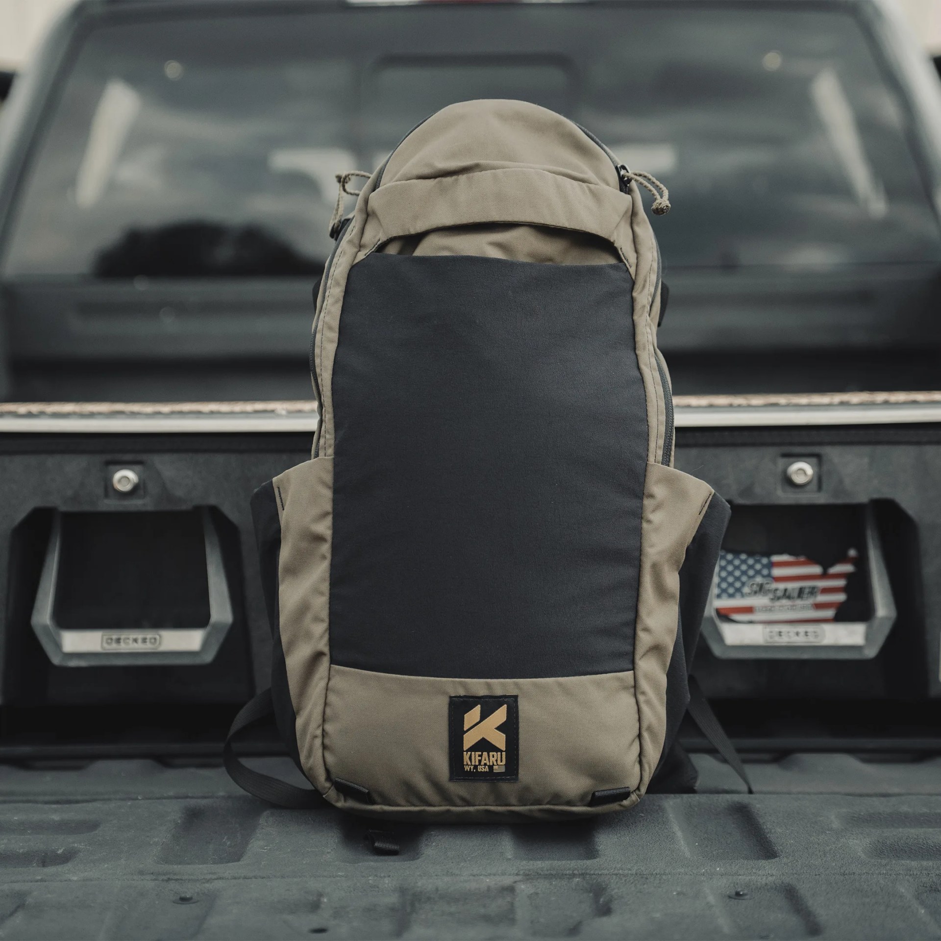Two-tone black and olive green Kifaru backpack standing upright on a truck bed with storage compartments in the background.