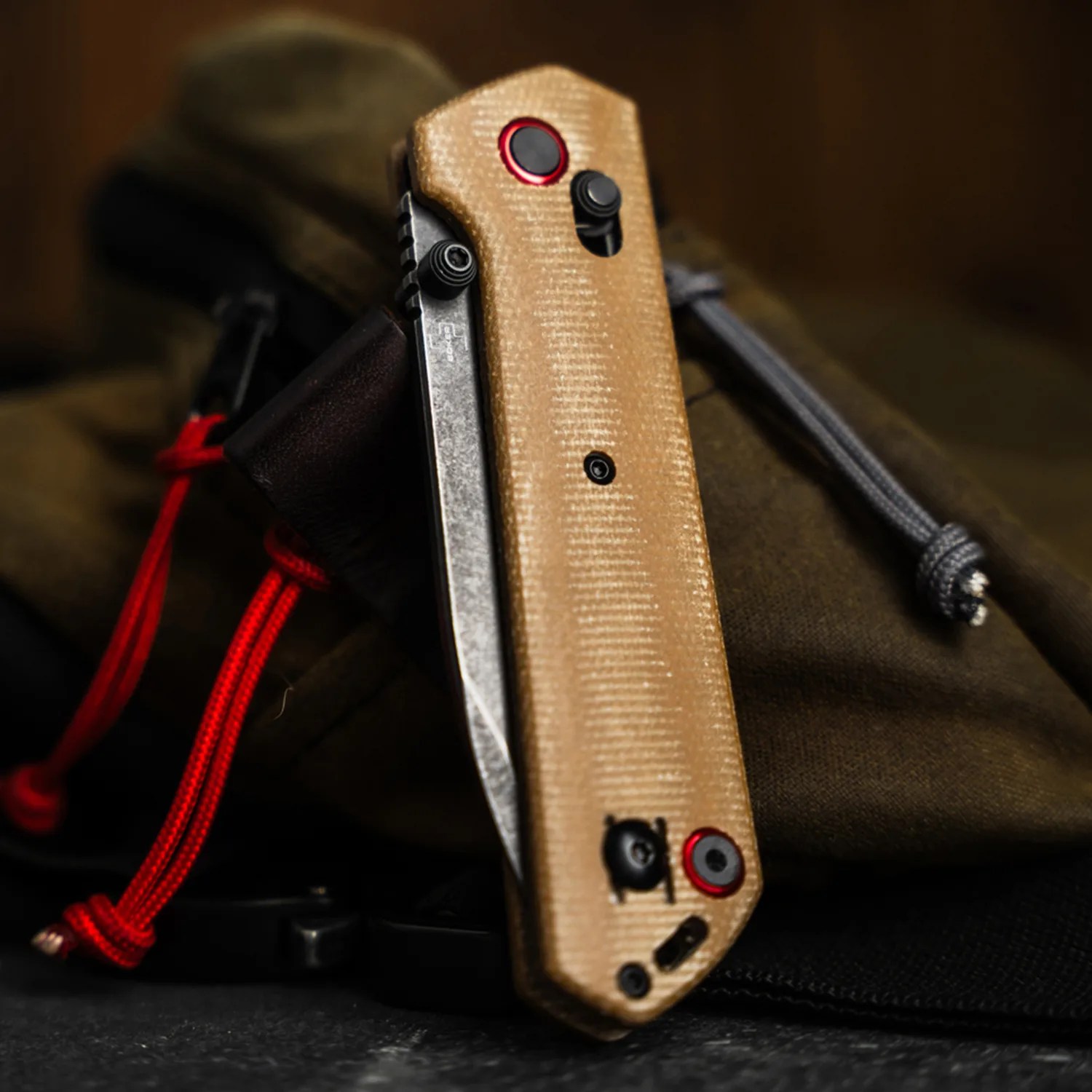 Folding knife with tan textured handle and black and red hardware resting on dark fabric with red and gray cords.