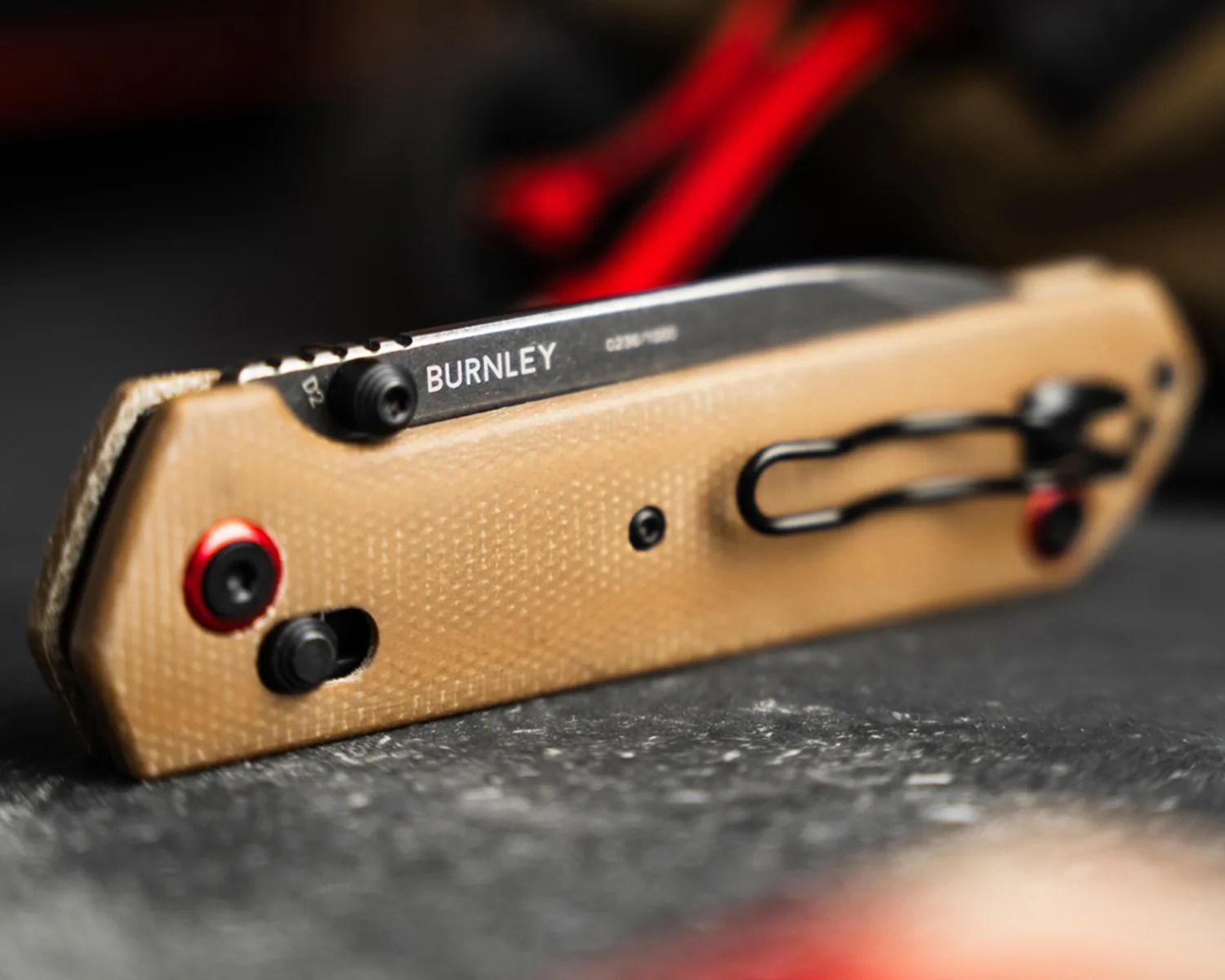Tan textured folding knife handle with black clip and red-accented screws on a dark surface.