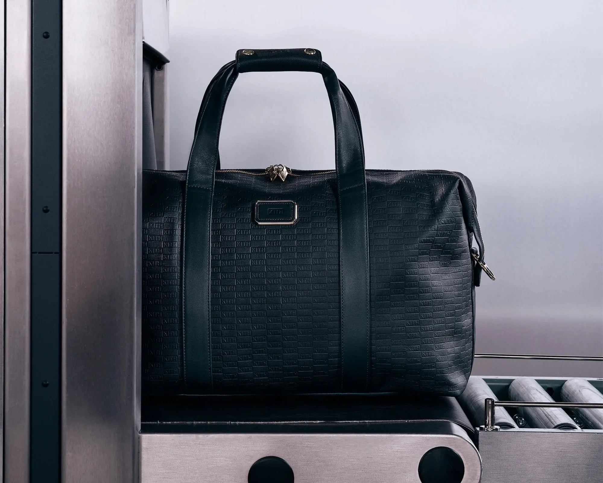 Black textured leather duffel bag with double handles on a metal conveyor belt.