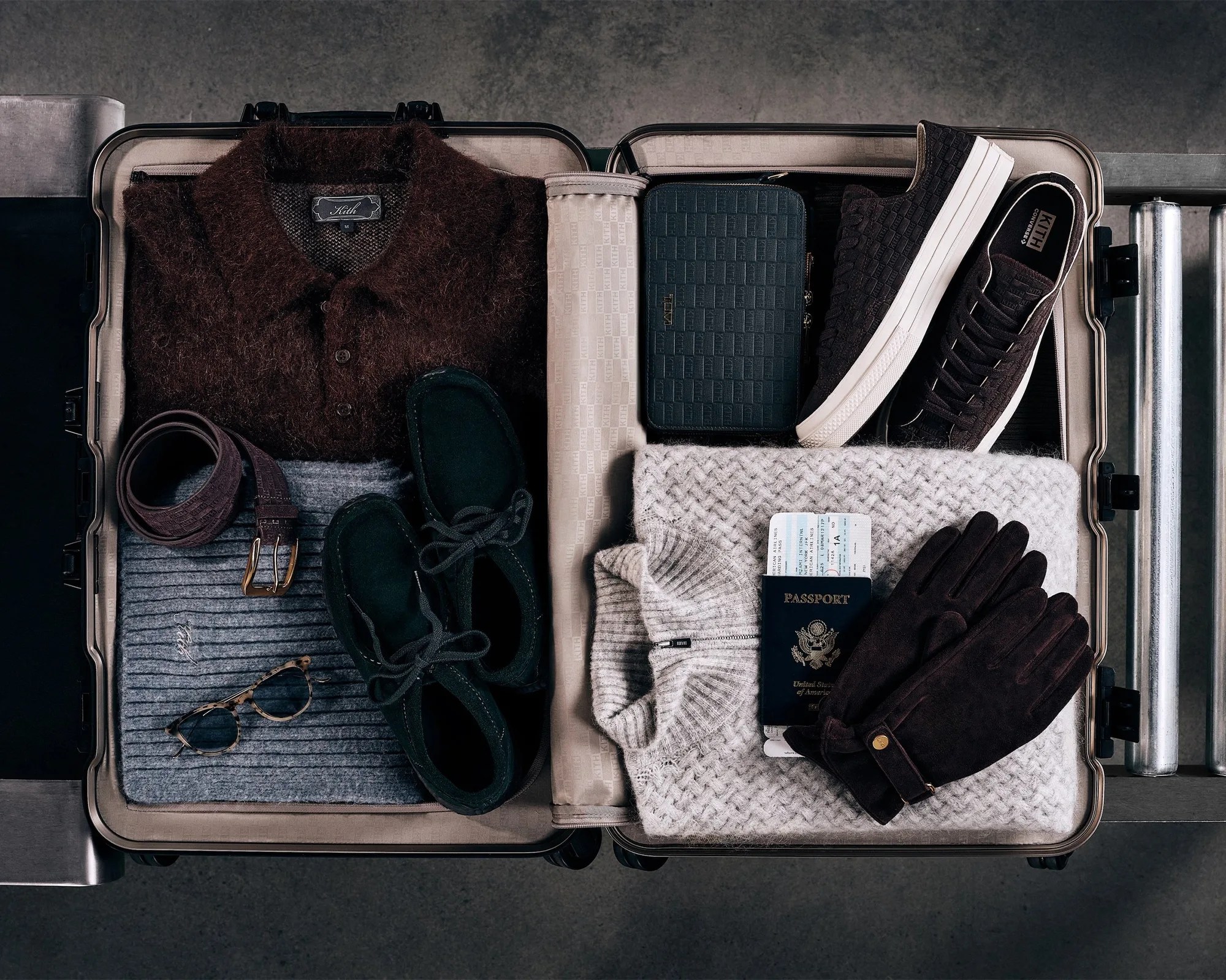 Open suitcase with folded brown and gray sweaters, black shoes, belt, tortoiseshell glasses, black wallet, black sneakers, gray knit sweater, passport, and black gloves.