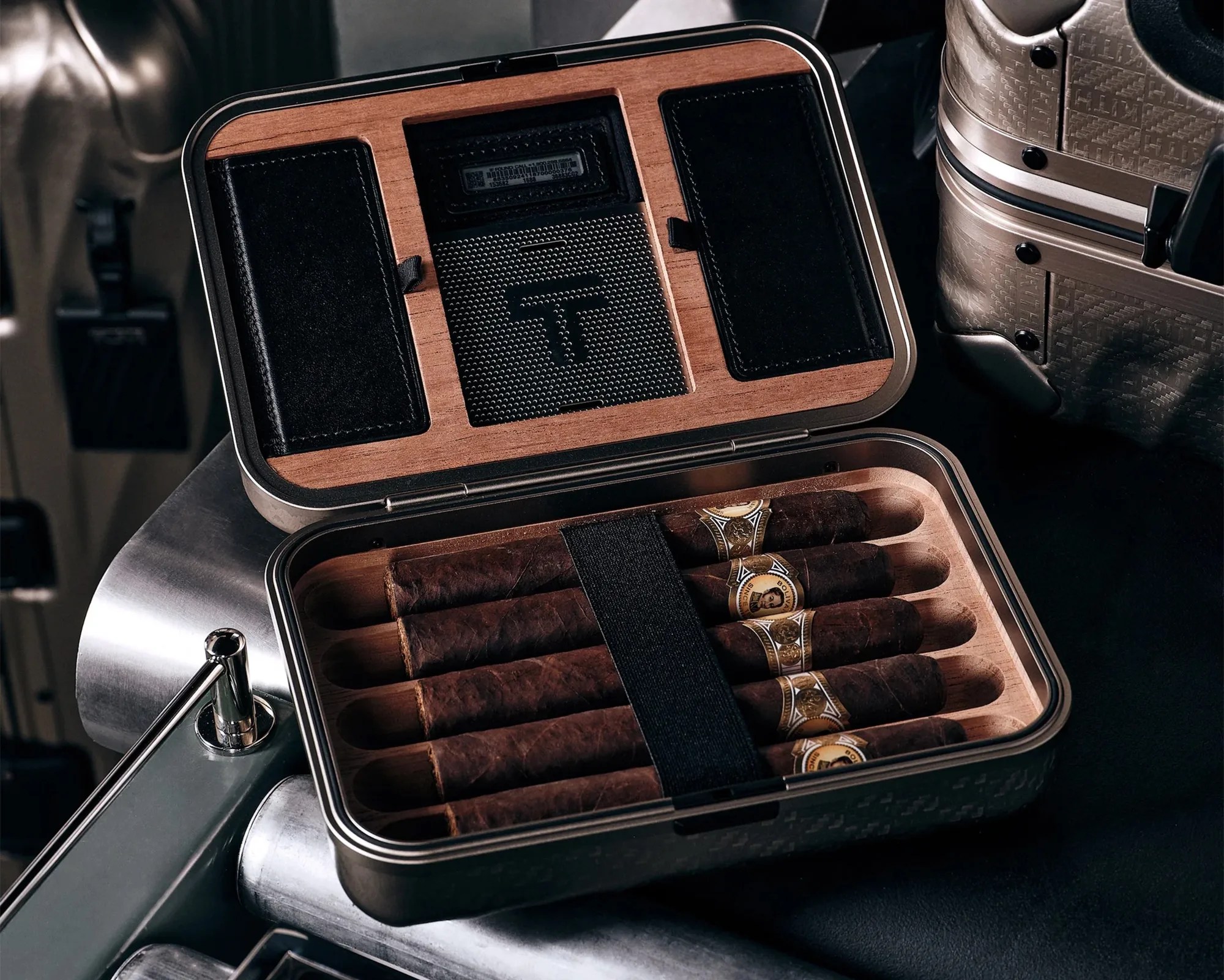 Open metal and wood cigar humidor case with five dark brown cigars secured by a black strap inside.