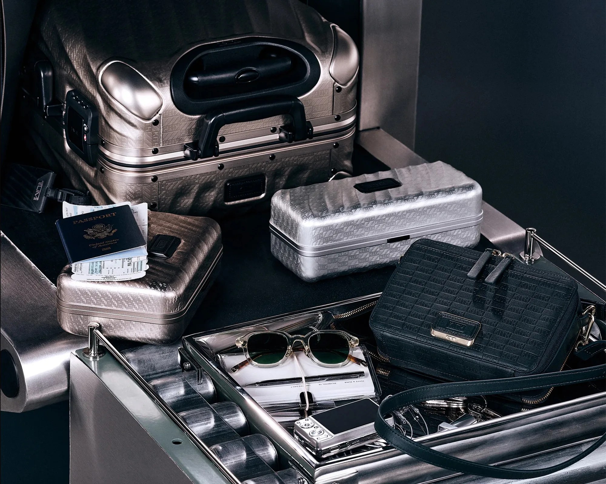 Set of metallic silver and black textured travel cases with a black leather crossbody bag, sunglasses, passport, and a digital camera on a luggage conveyor belt.