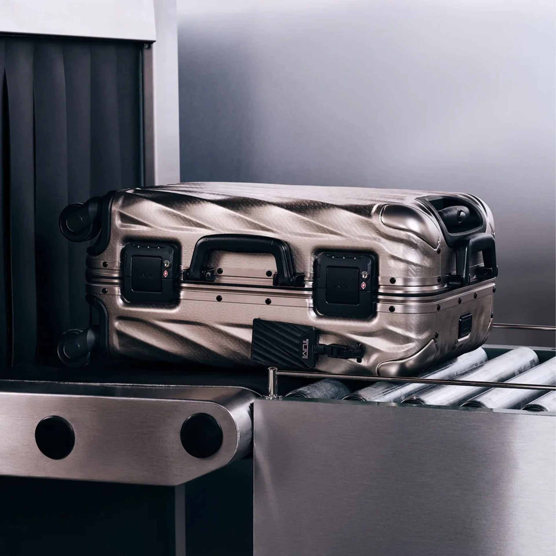 Metallic silver hard-shell suitcase with black handles and wheels on an airport conveyor belt.