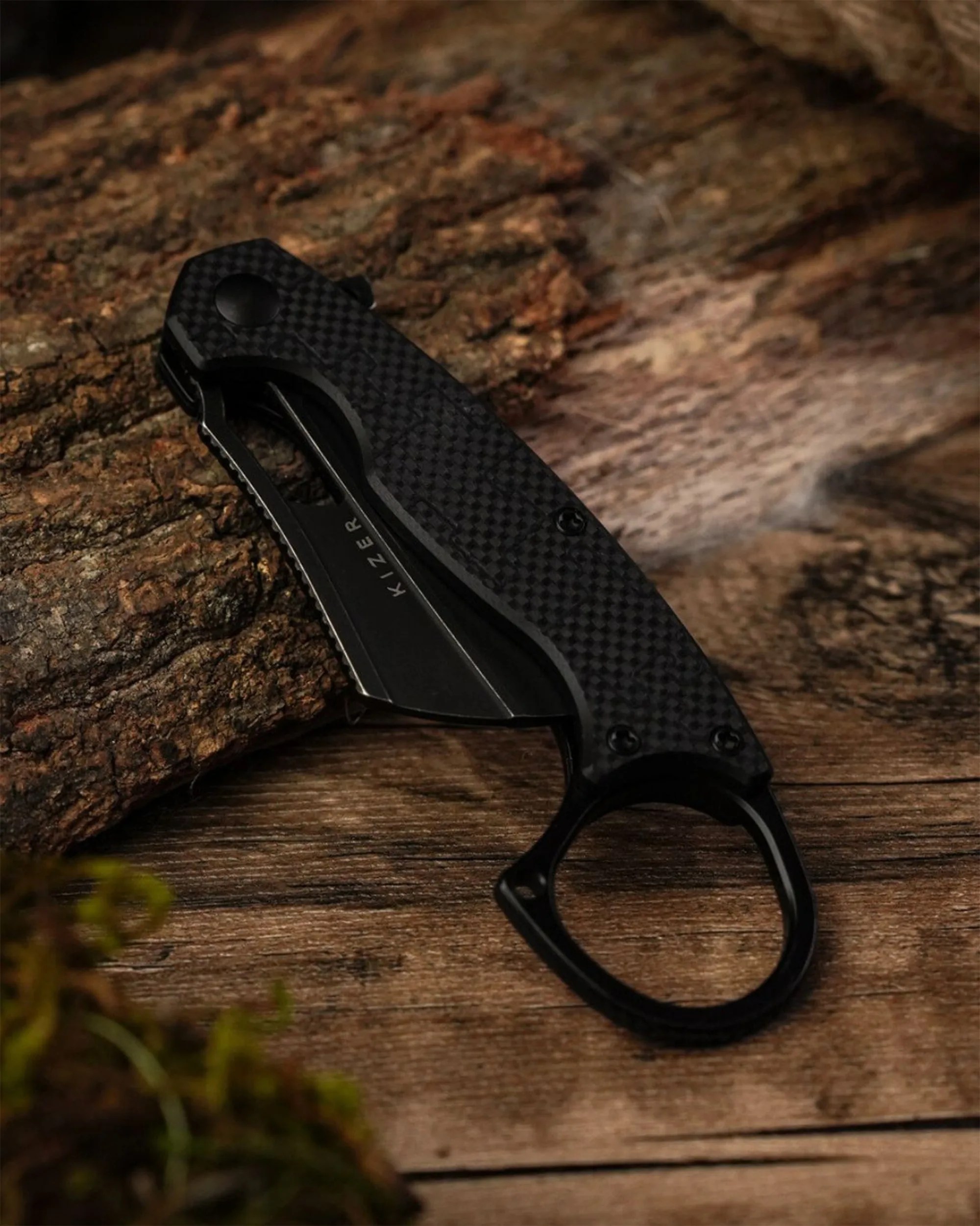 Black folding knife with textured handle and finger ring, resting on a wooden surface near bark.
