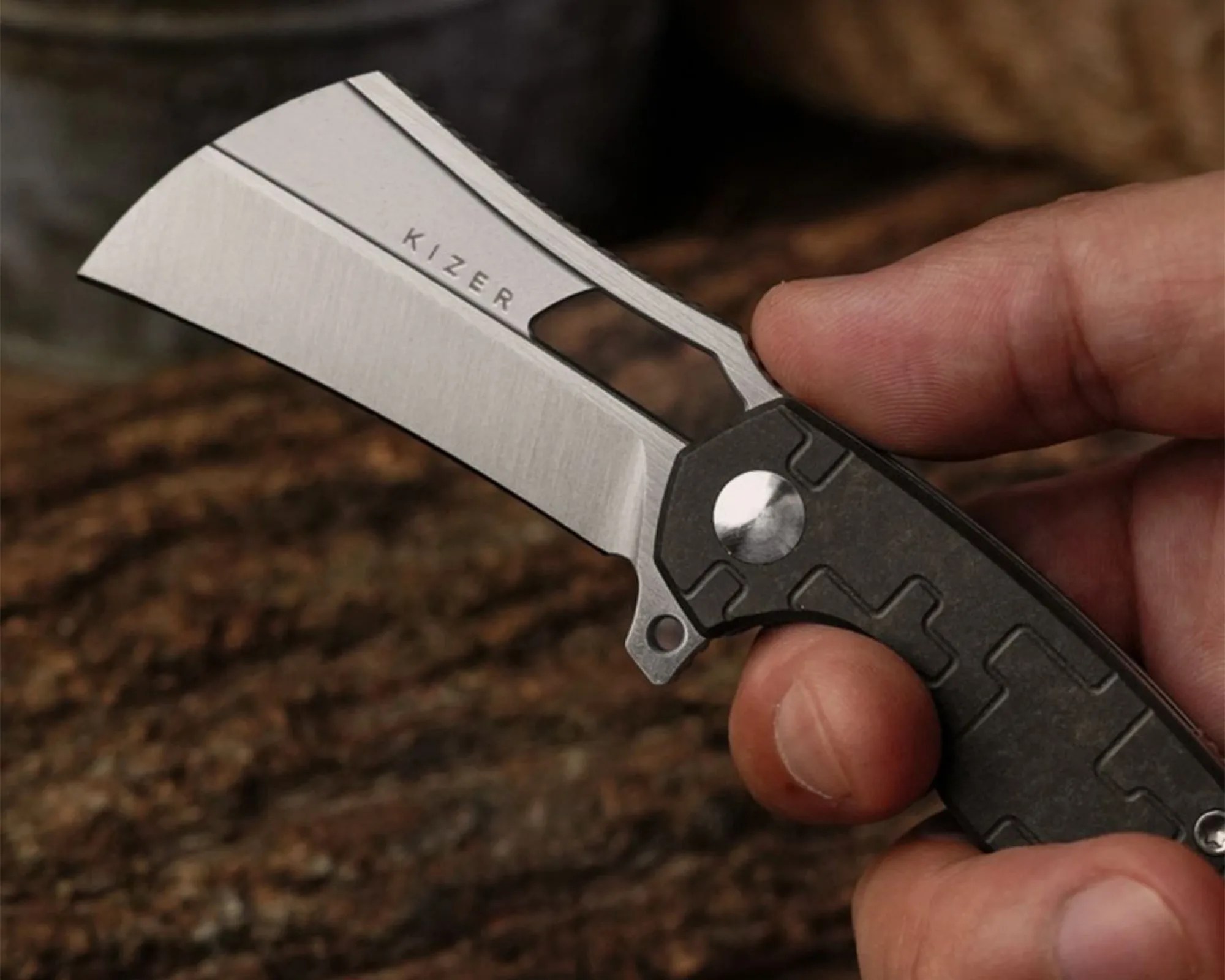 Hand holding a Kizer folding knife with a stonewashed cleaver-style blade and textured dark handle.