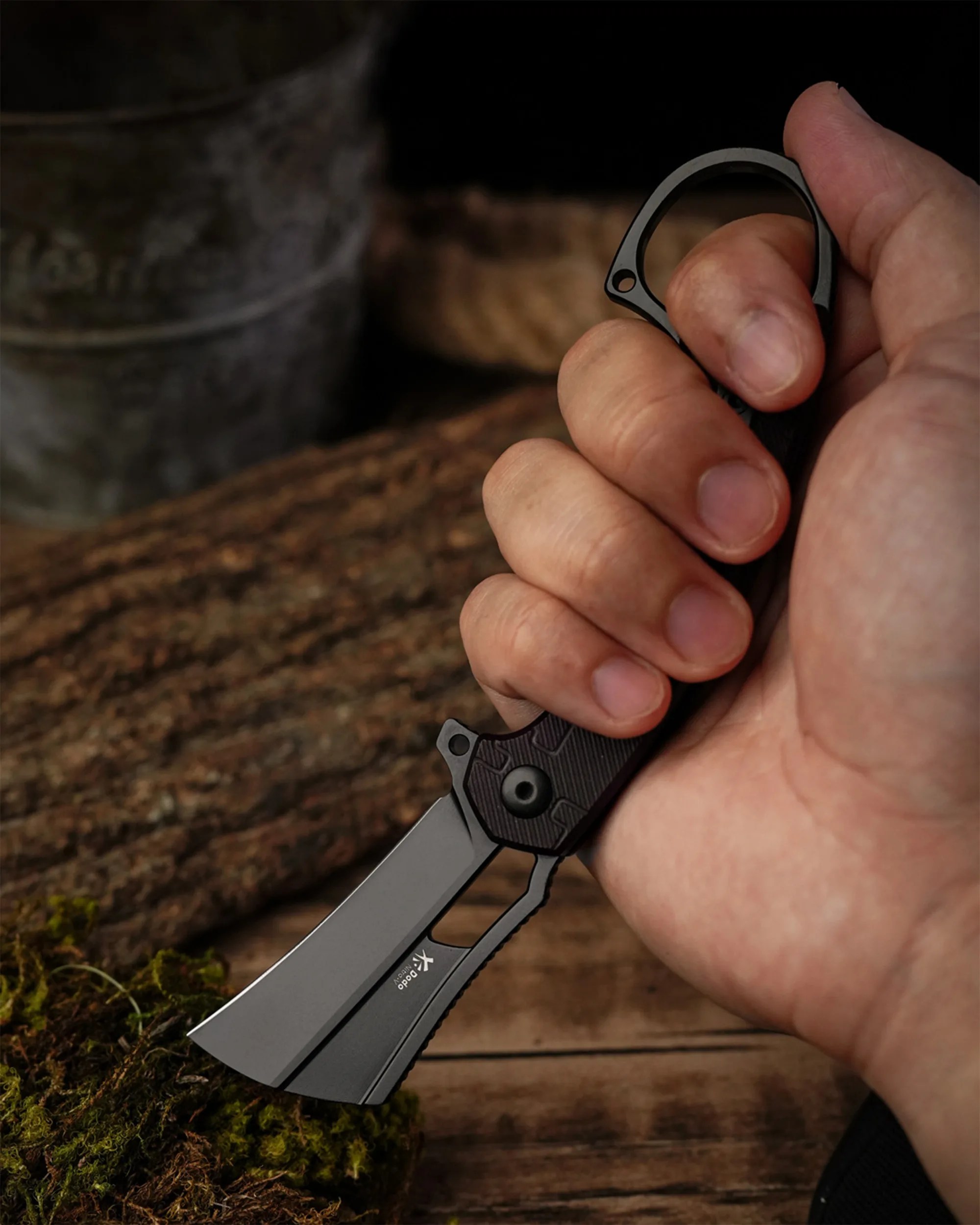 Hand holding a black folding knife with a cleaver-style blade over moss and wooden surface.