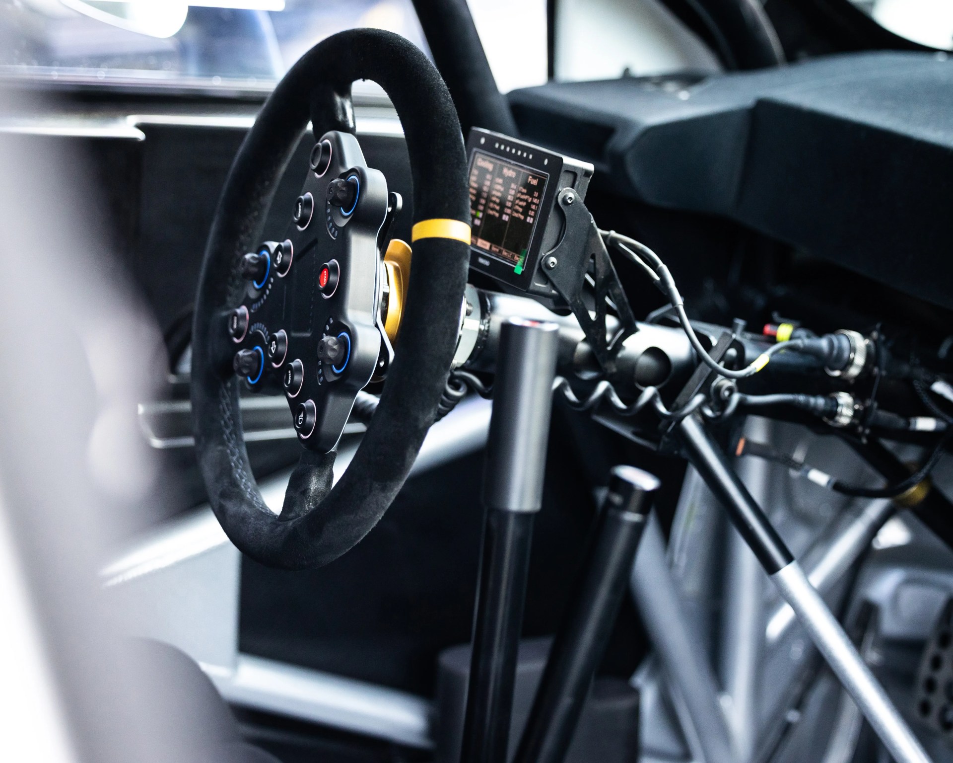 Black suede racing steering wheel with multiple control buttons and a digital display inside a race car cockpit.