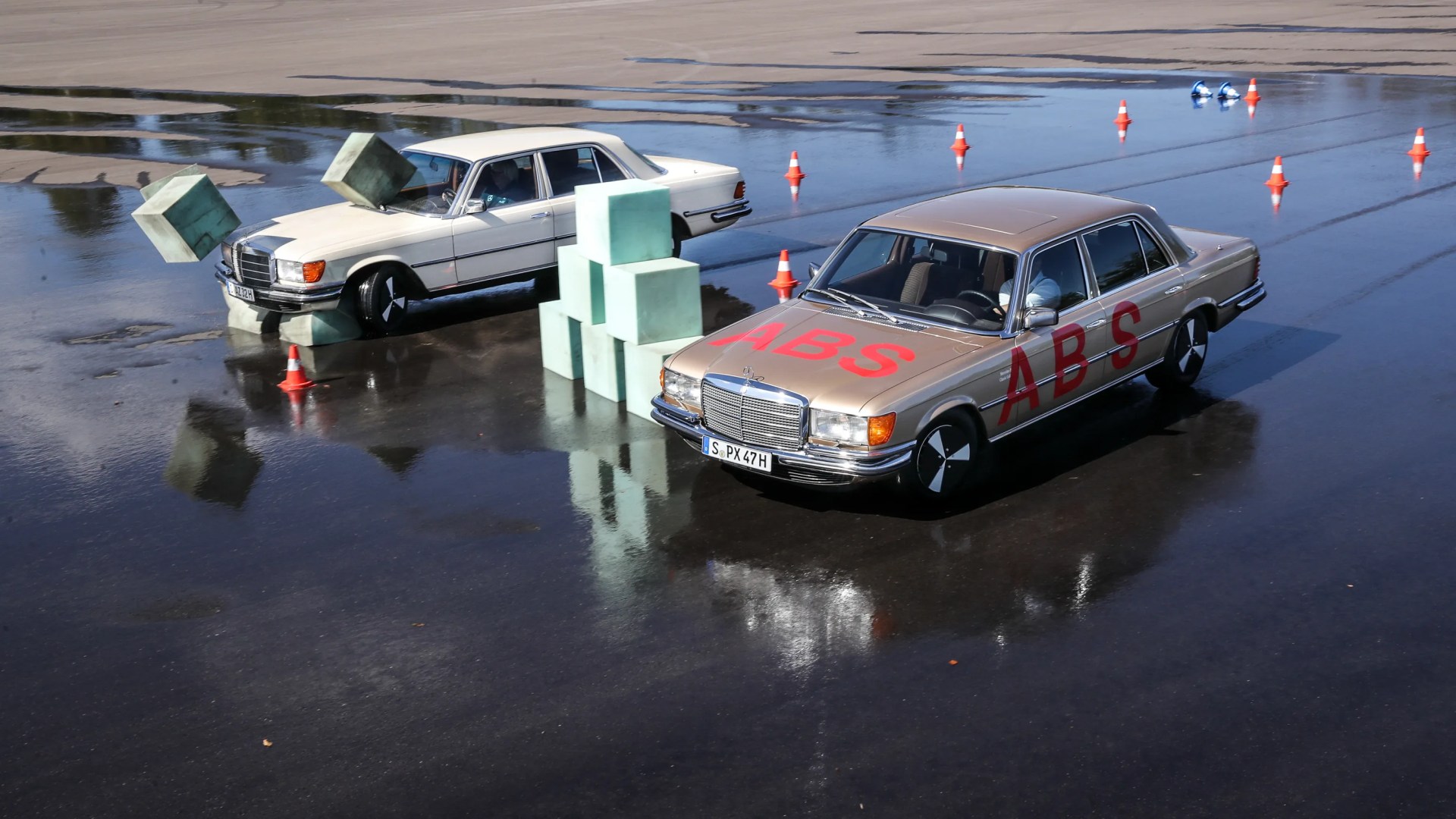 Two vintage Mercedes sedans on a wet asphalt surface, one with "ABS" in red letters and cones around, with green foam cubes stacked and falling.