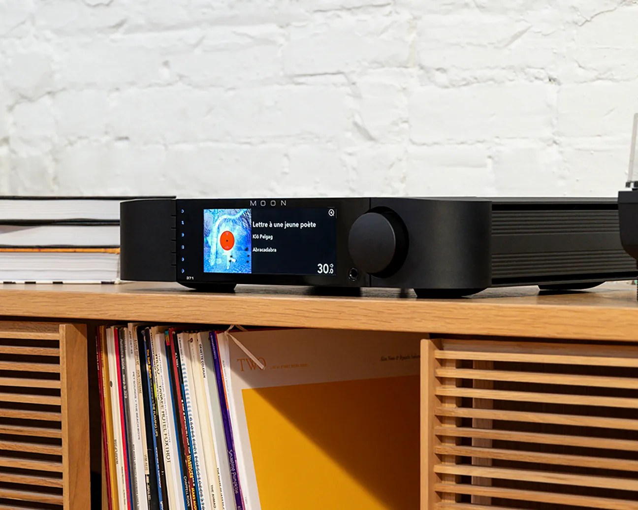 Black MOON audio device with digital display showing album art and track info, placed on a wooden shelf with vinyl records.