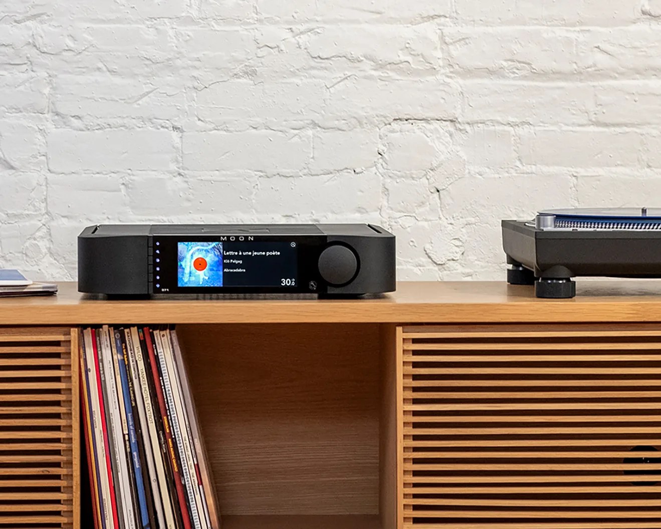 Black MOON audio player with digital display showing album art and track info on a wooden shelf next to a turntable.